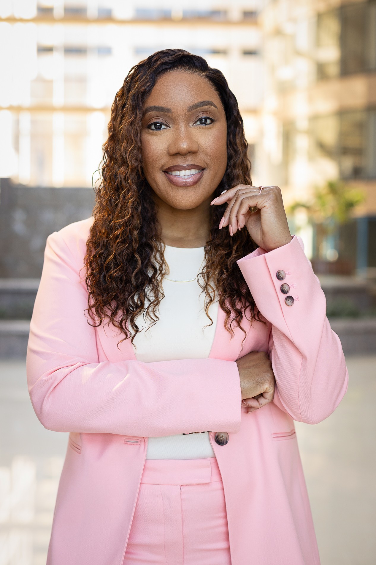Woman with curly hair wearing a pink blazer and white top, smiling outdoors with buildings in background.