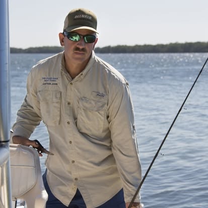 Man in beige shirt, sunglasses, and cap fishing on a boat with water and distant shoreline in background.