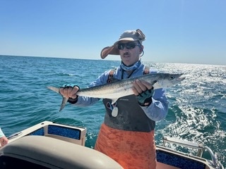 Person wearing sunglasses, hat, and fishing gear holding a fish on a boat with ocean and sky in background.