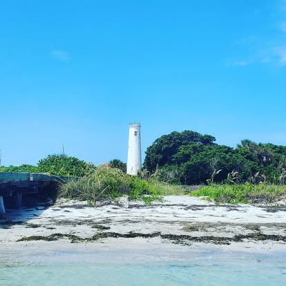 Lighthouse on a grassy hill with trees, sandy beach, and blue sky.