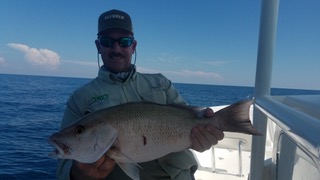 Person holding a large fish on a boat with ocean background, wearing sunglasses and a cap.