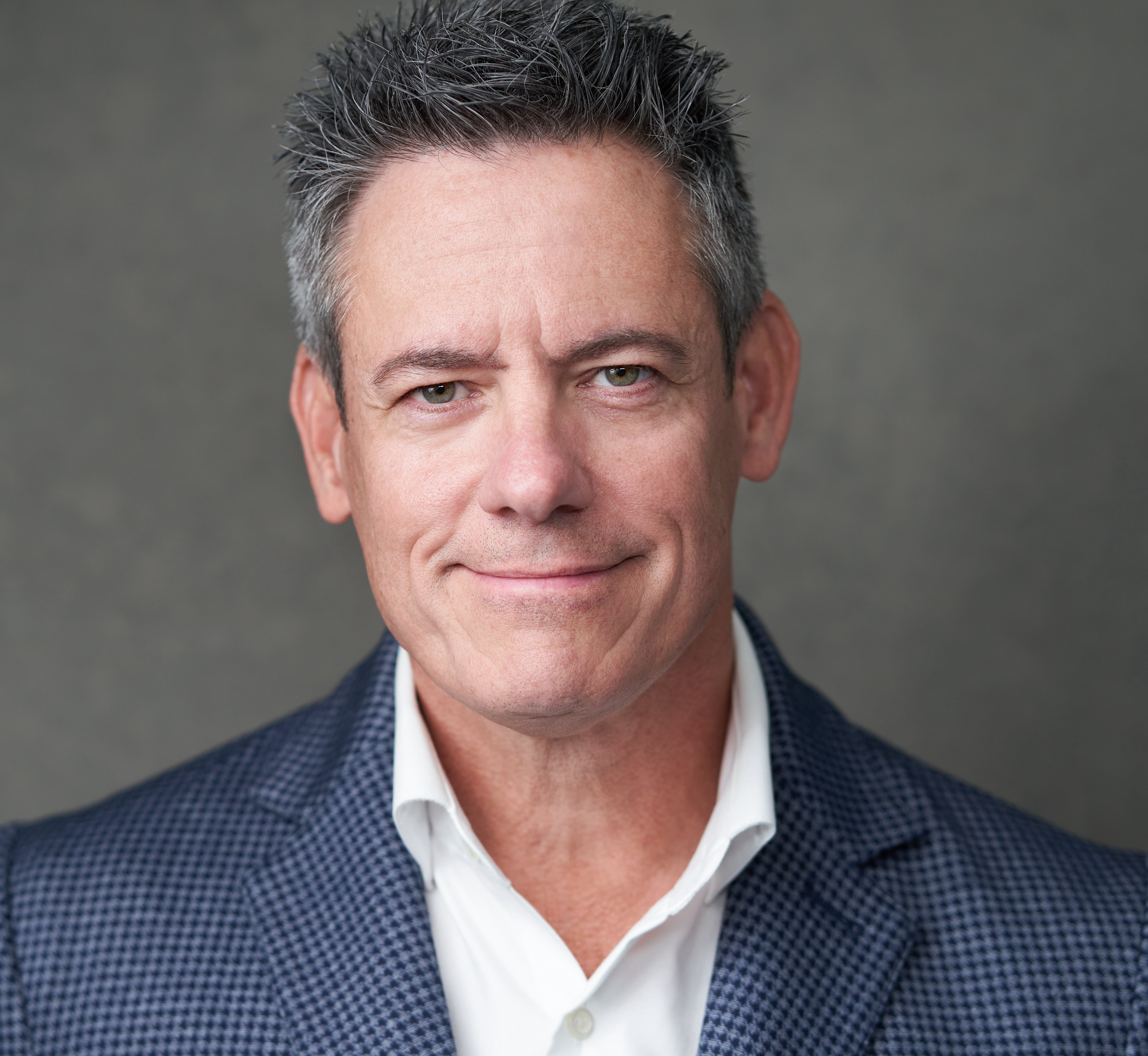 Man with short, dark hair and a slight smile, wearing a dark checkered blazer and white shirt, against a plain background.