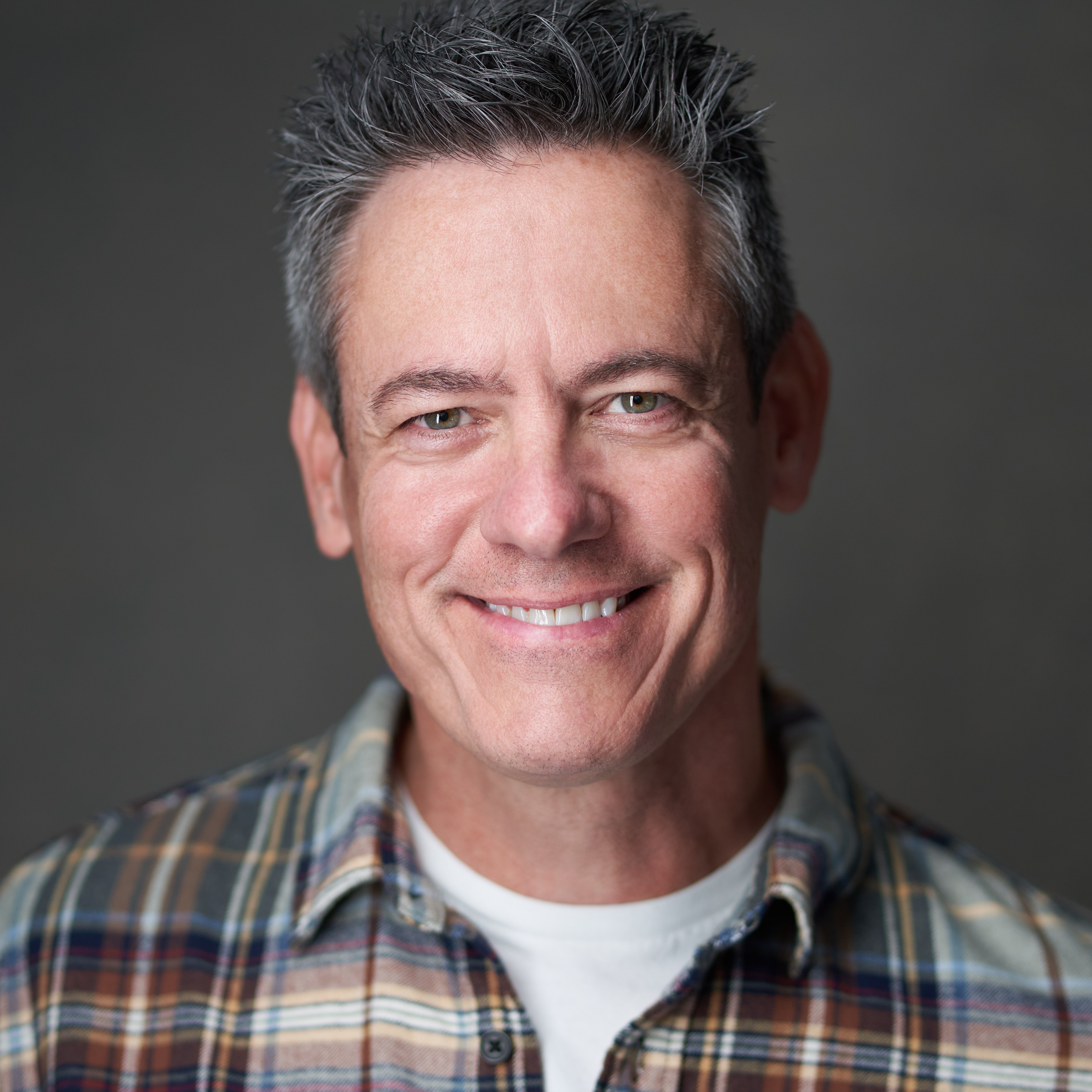Smiling man with short, gray hair wearing a plaid shirt and white undershirt, facing forward.