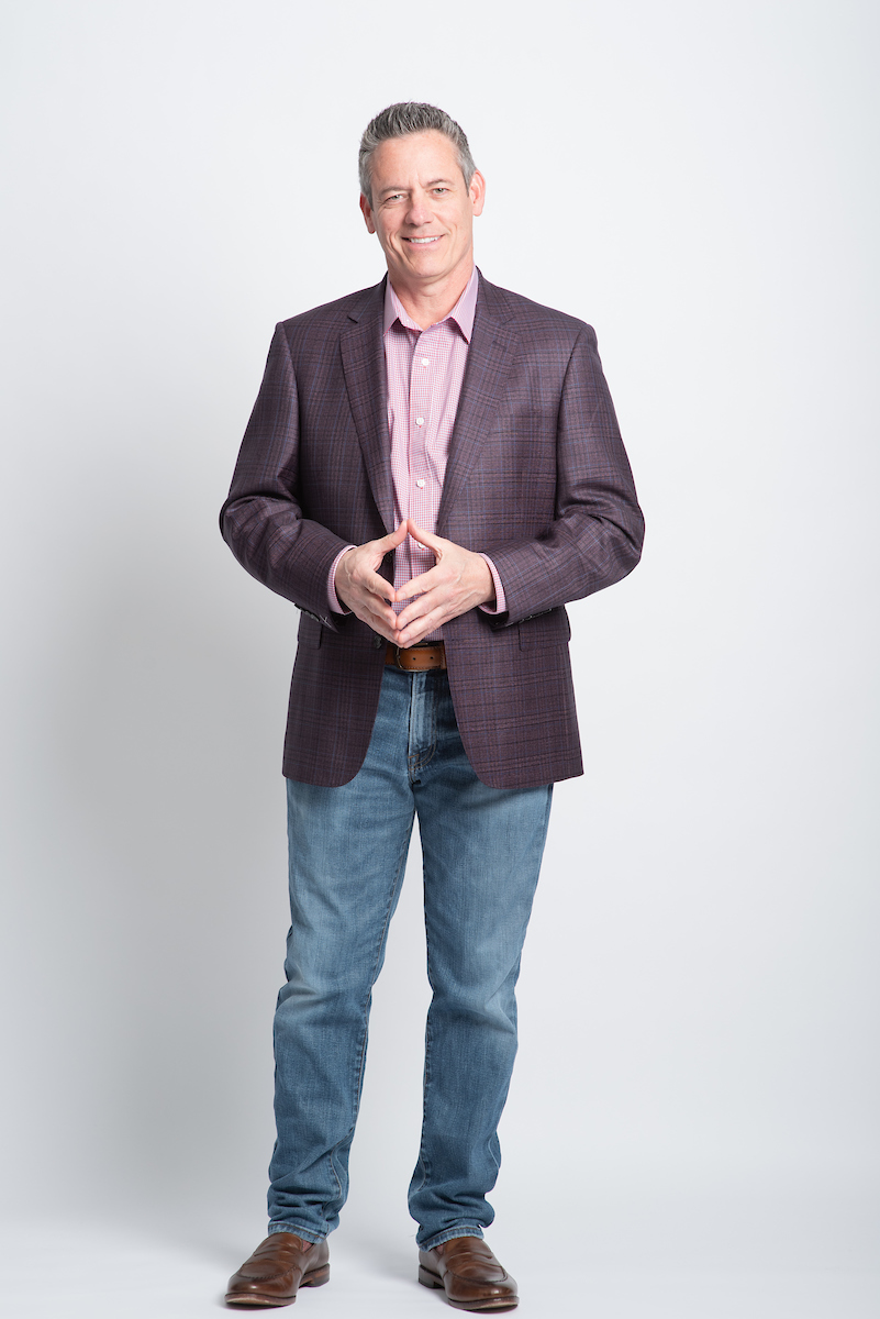 Man in blazer and jeans standing with hands clasped, smiling, against a plain white background.