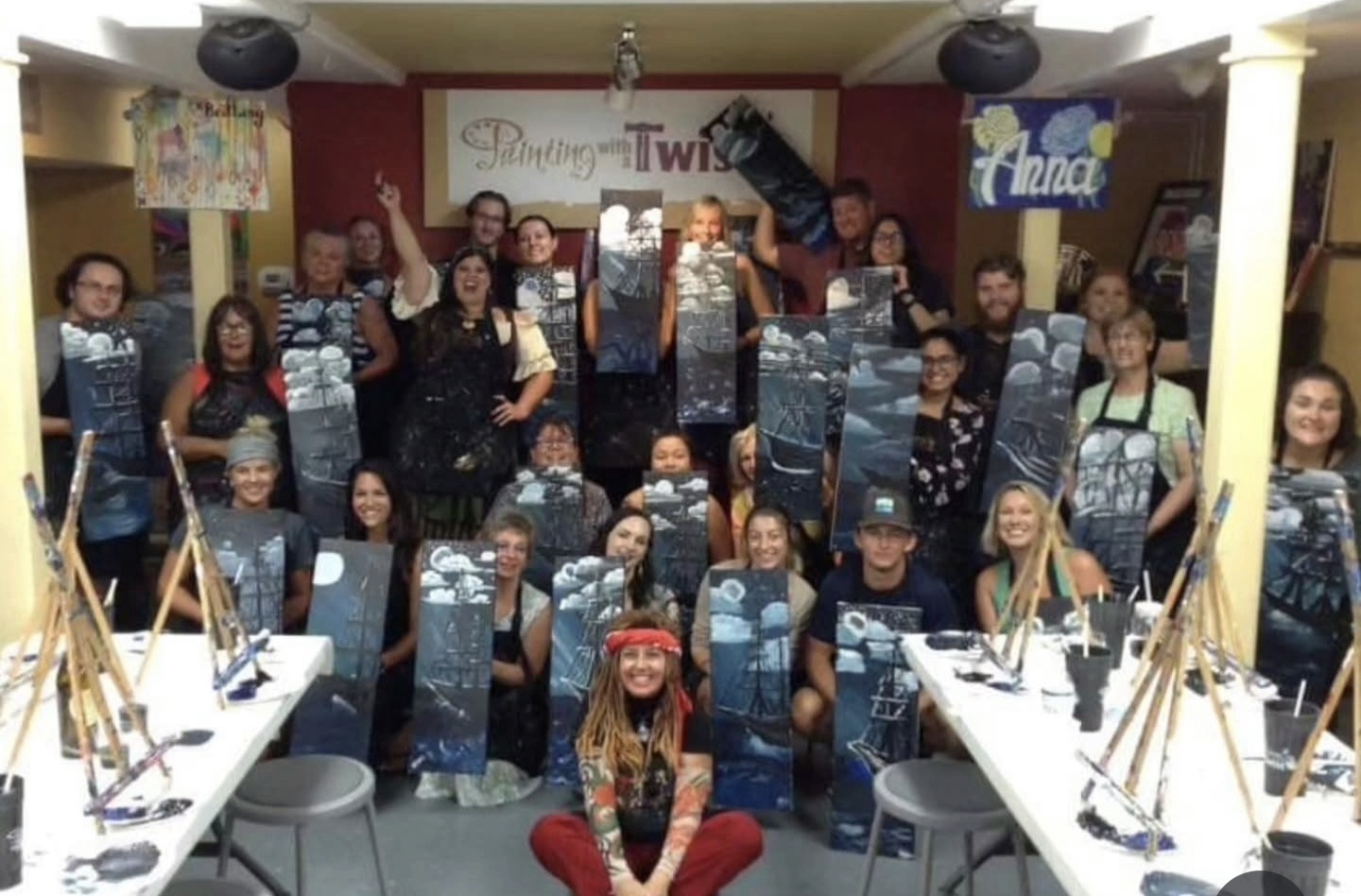 Group of people in a room holding up paintings, with tables and art supplies visible, celebrating art event.