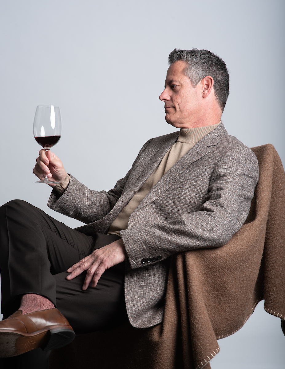 Man in a gray blazer and beige sweater sitting in a chair, holding a glass of red wine, against a plain background.