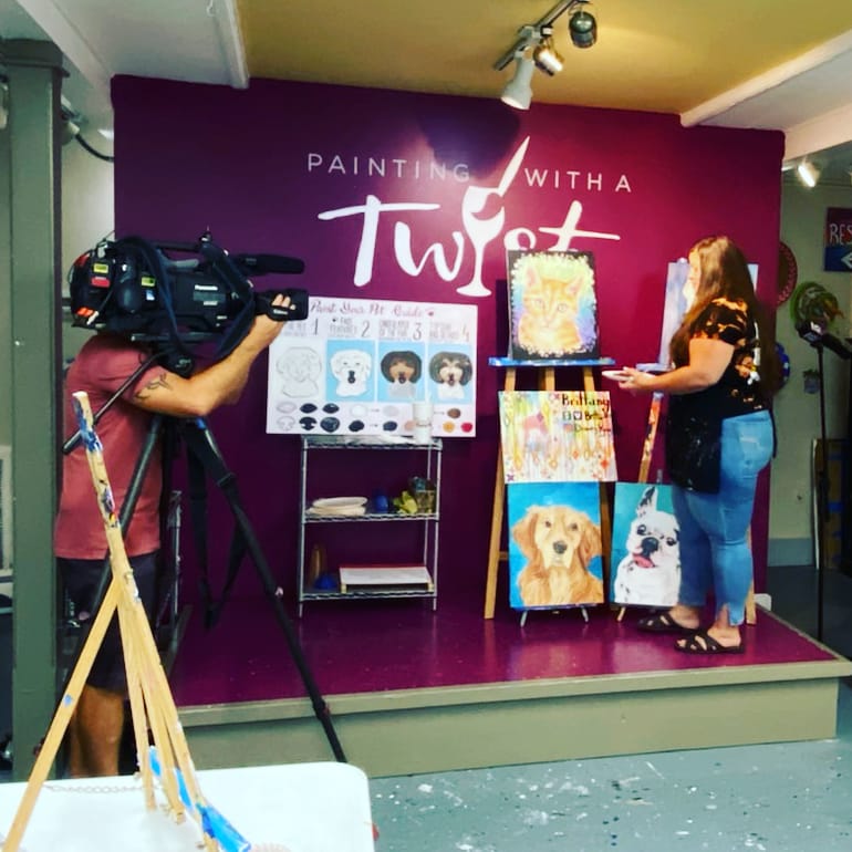 Person filming a woman painting dog portraits on a small stage with a purple wall background, text, and paintings displayed. Word: 'Painting with a Twist'.
