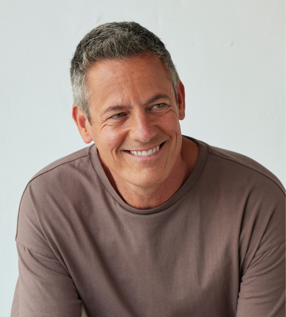 Smiling middle-aged man with short gray hair wearing a brown shirt, facing forward, against a plain light background.