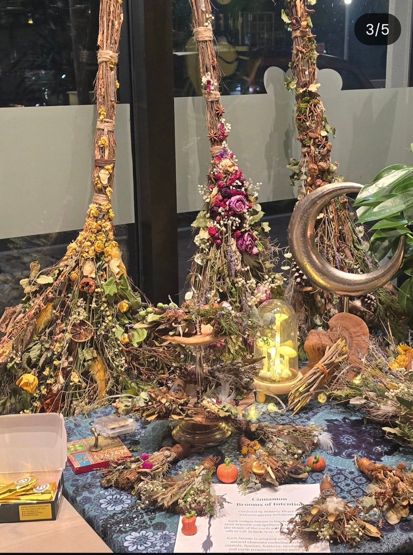 Display of floral arrangements with dried flowers, candles, and decorative items on a table, viewed through a window.