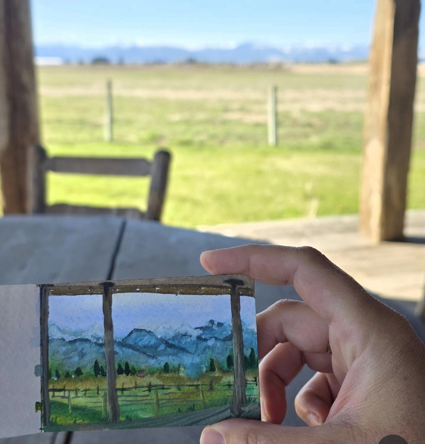 Person holding a small watercolor painting of mountains and trees, outdoors with a wooden structure and open landscape in background.