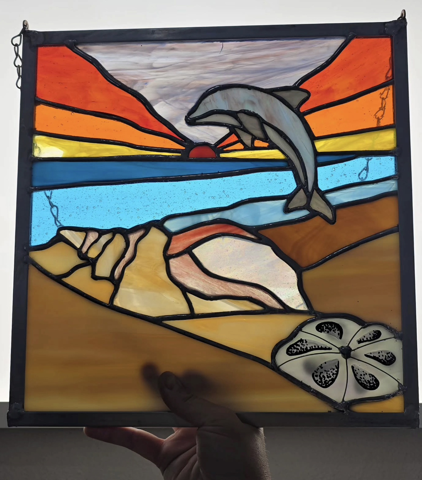 Colorful stained glass window depicting a dolphin jumping over a sunset, with land and water below, and a flower in the corner.