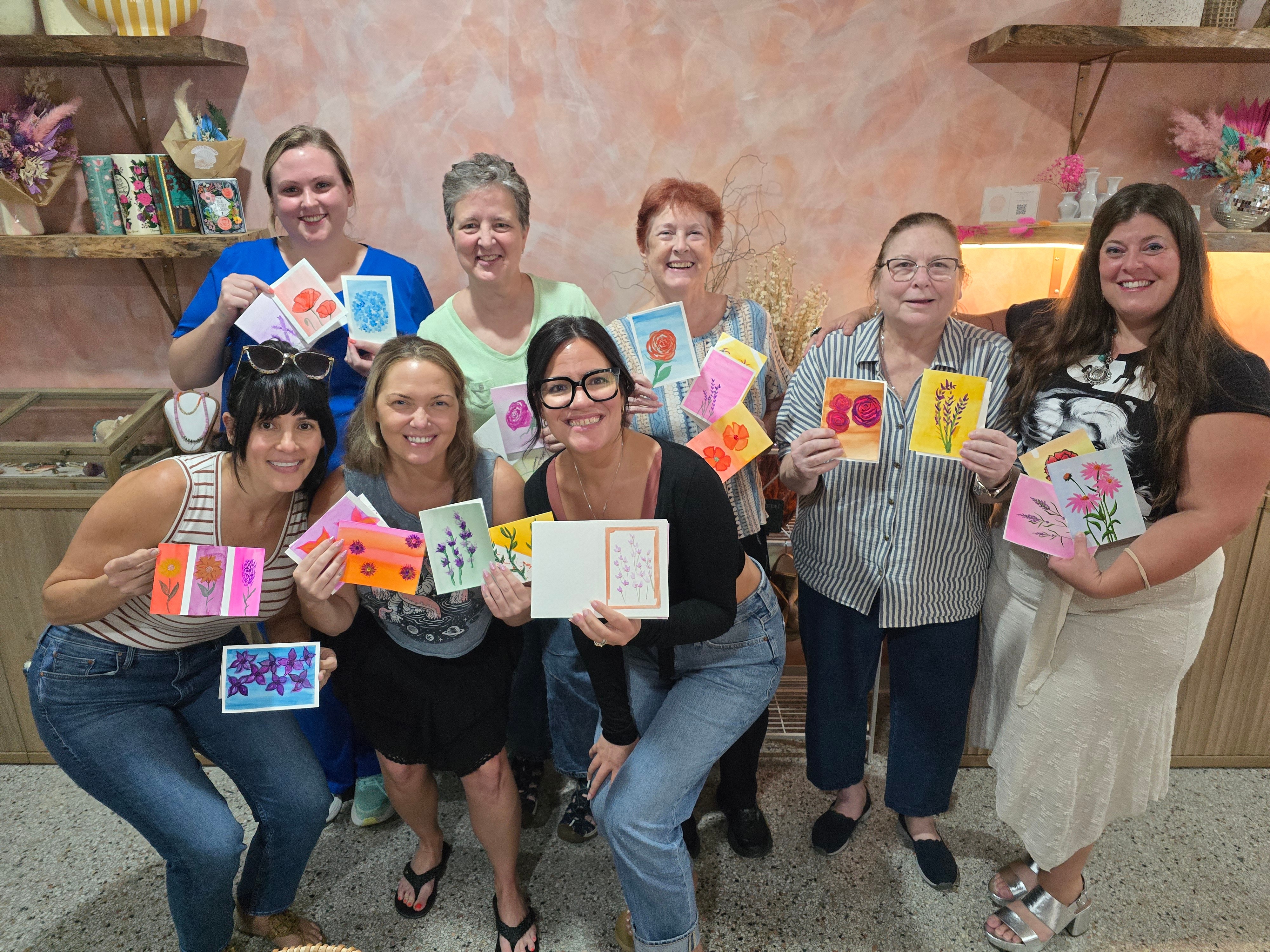 Group of eight women holding colorful handmade cards, smiling indoors with shelves and pink flowers in background.