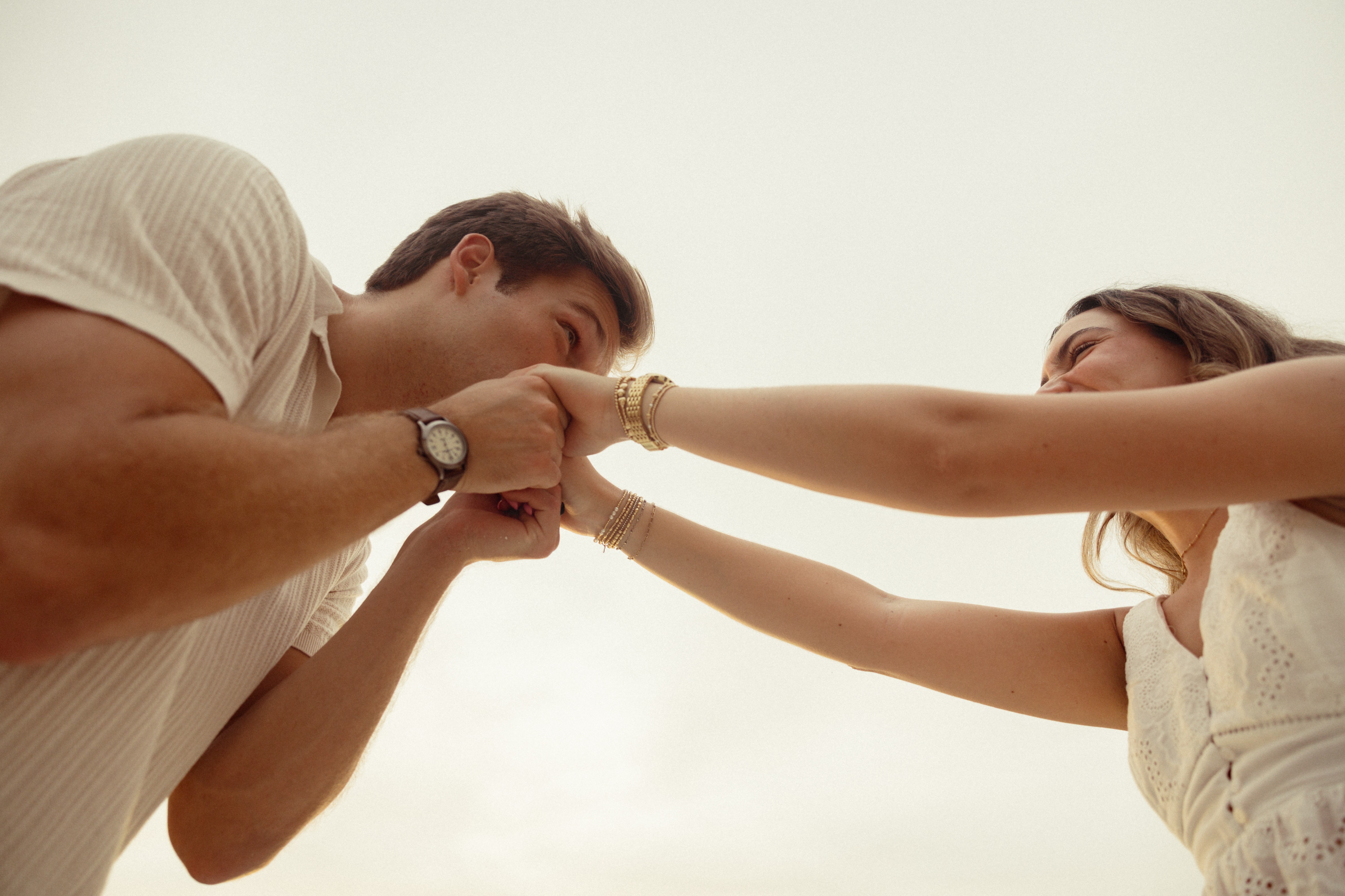Two people hold hands and kiss each other outdoors against a cloudy sky, with one person's face partially visible.