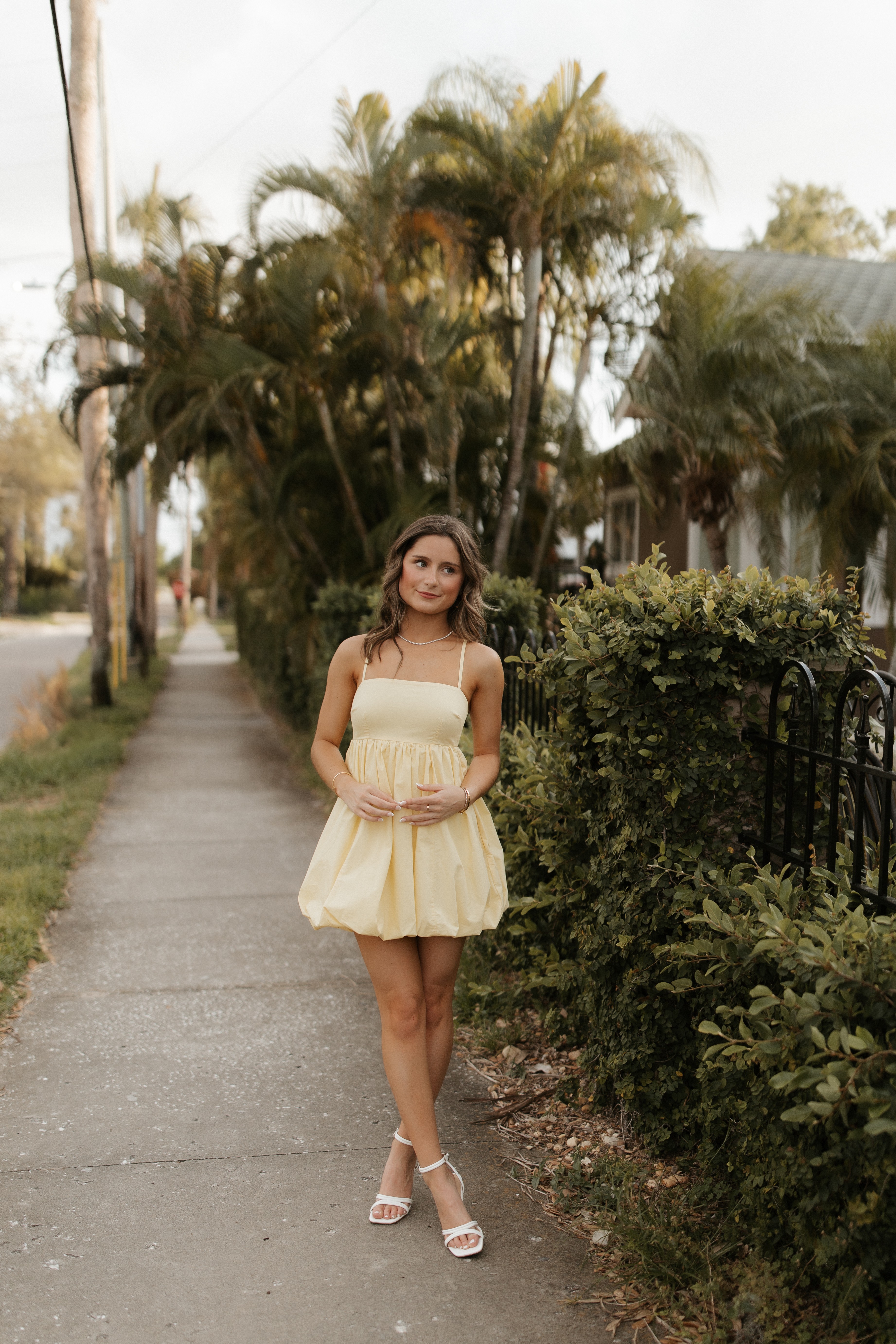 Young woman in yellow dress standing on sidewalk with palm trees and bushes in background.