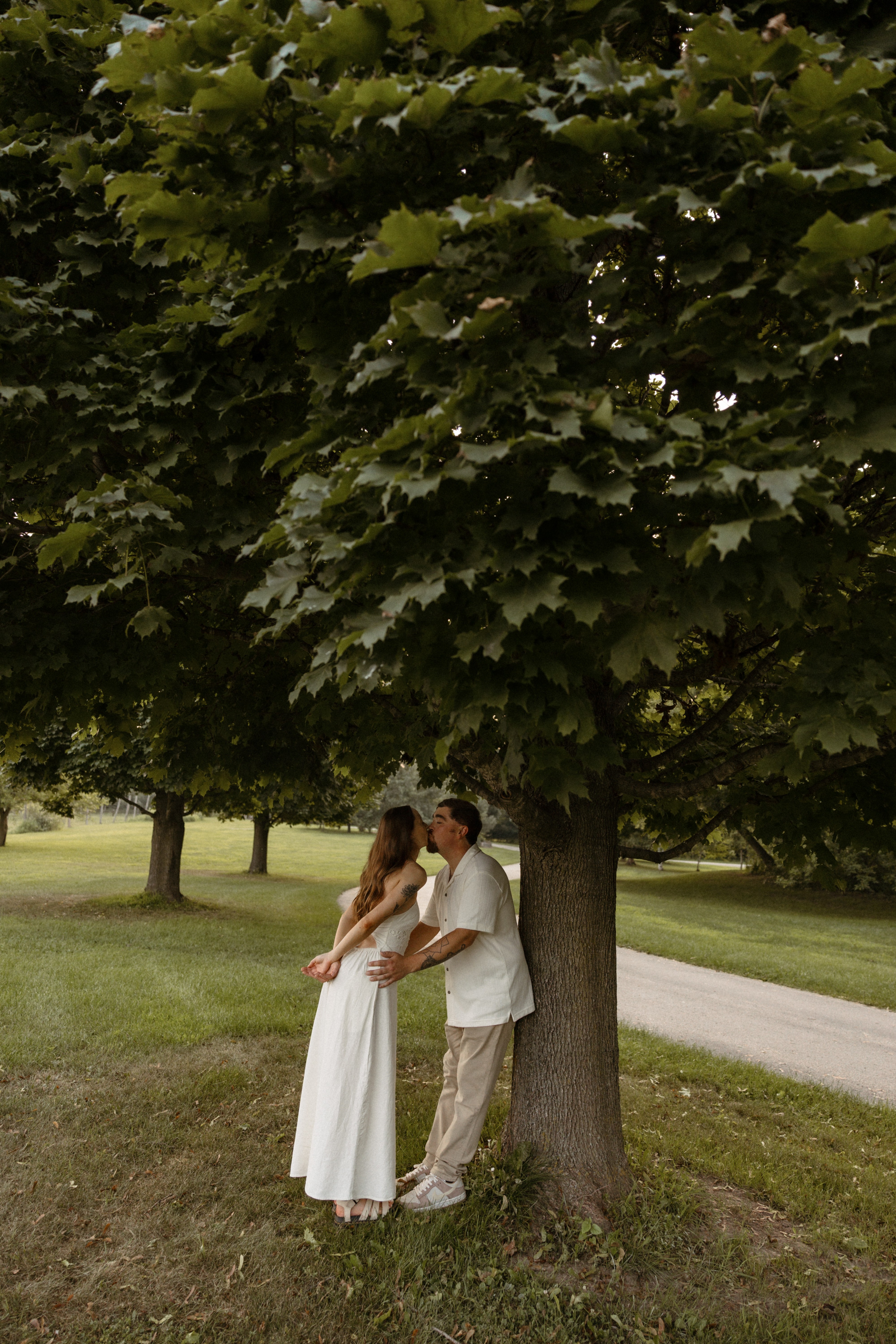Couple standing under a large leafy tree in a park, facing each other, with a grassy area and a path nearby.