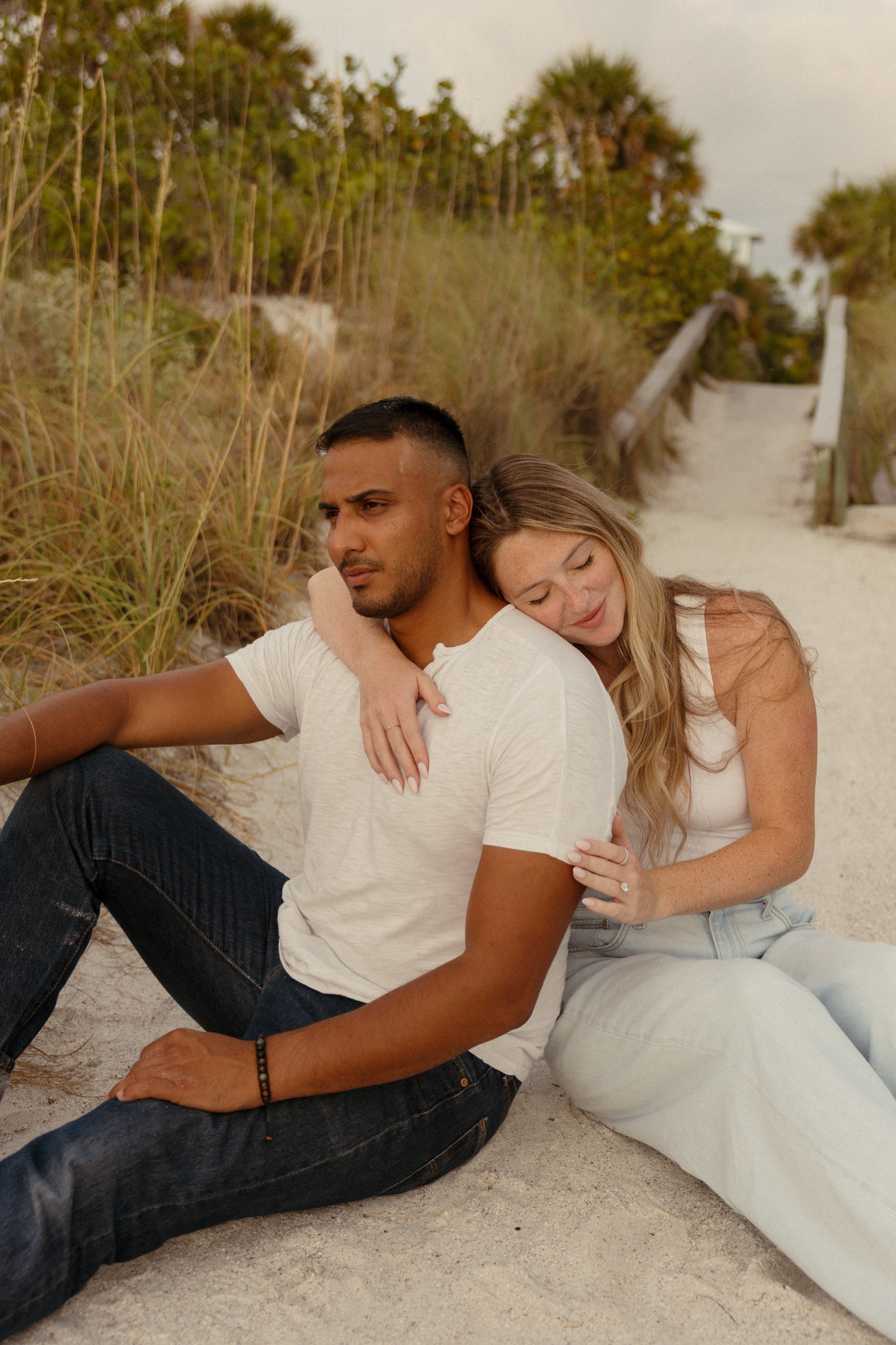 A man and woman sit on sand, with the woman resting her head on the man's shoulder, outdoors with trees and a slide in background.