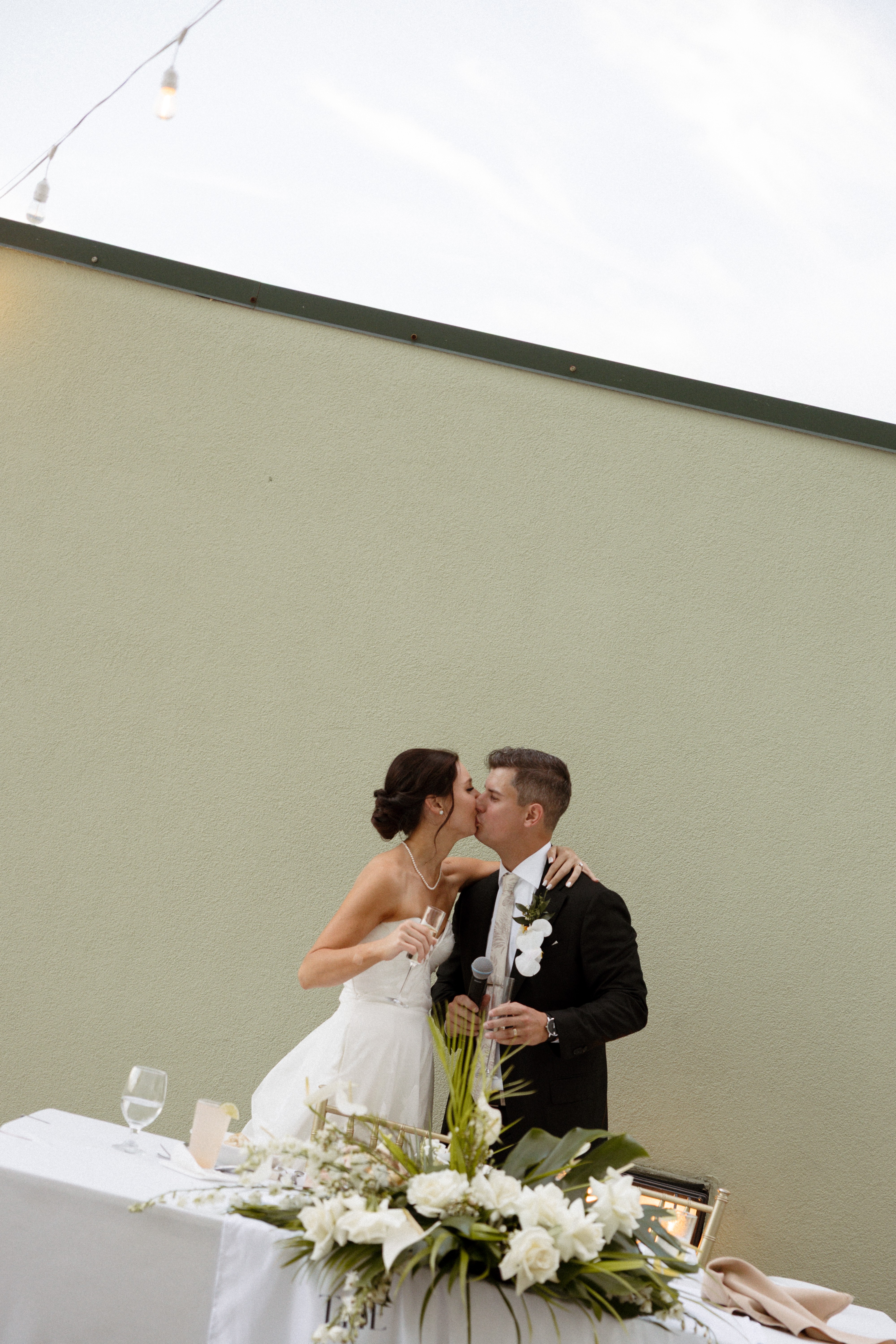 Bride and groom stand close together, holding flowers, in front of a plain wall during a wedding ceremony.