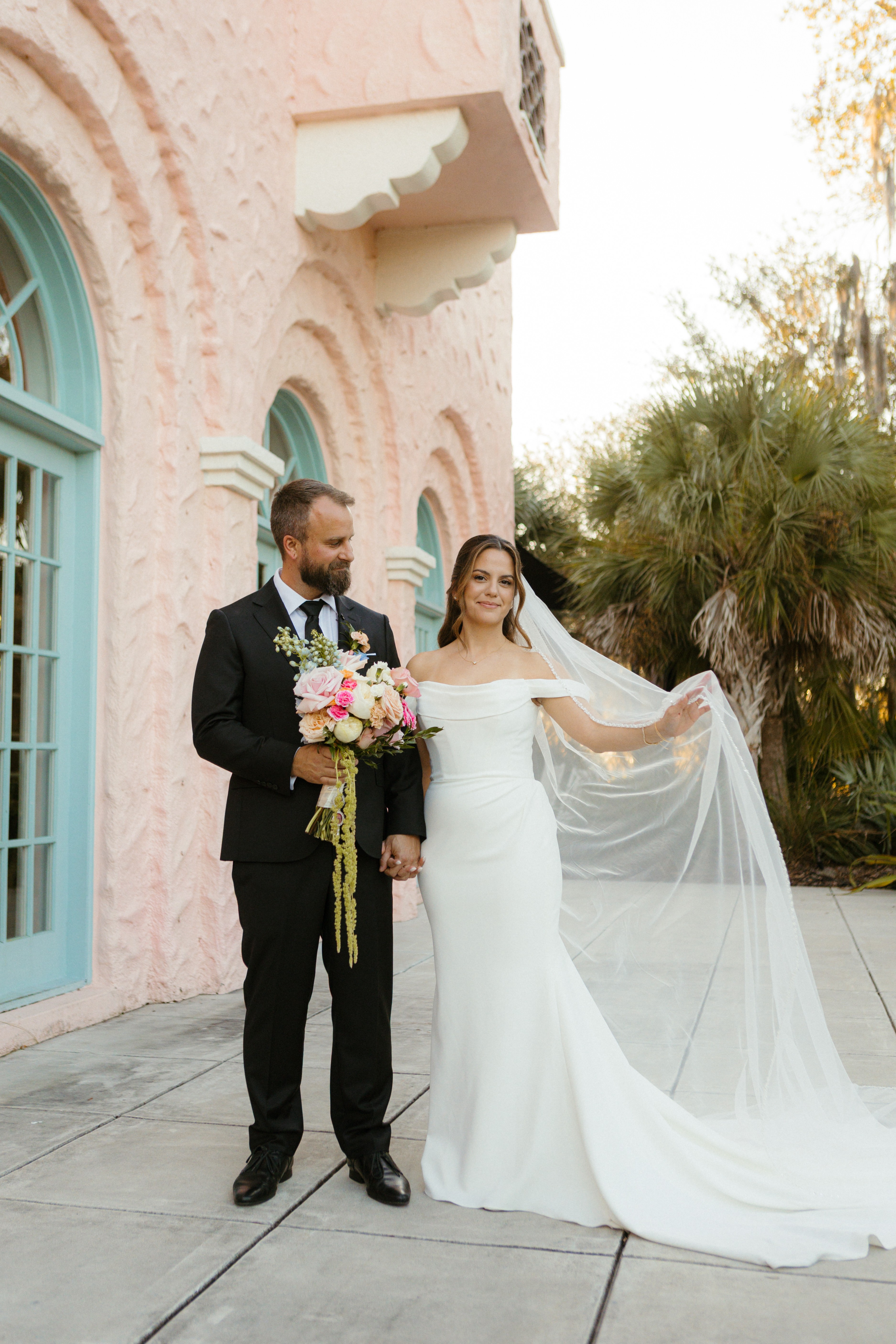 Bride and groom standing outside a pink building with arched windows, bride holding her veil, groom holding a bouquet of flowers.