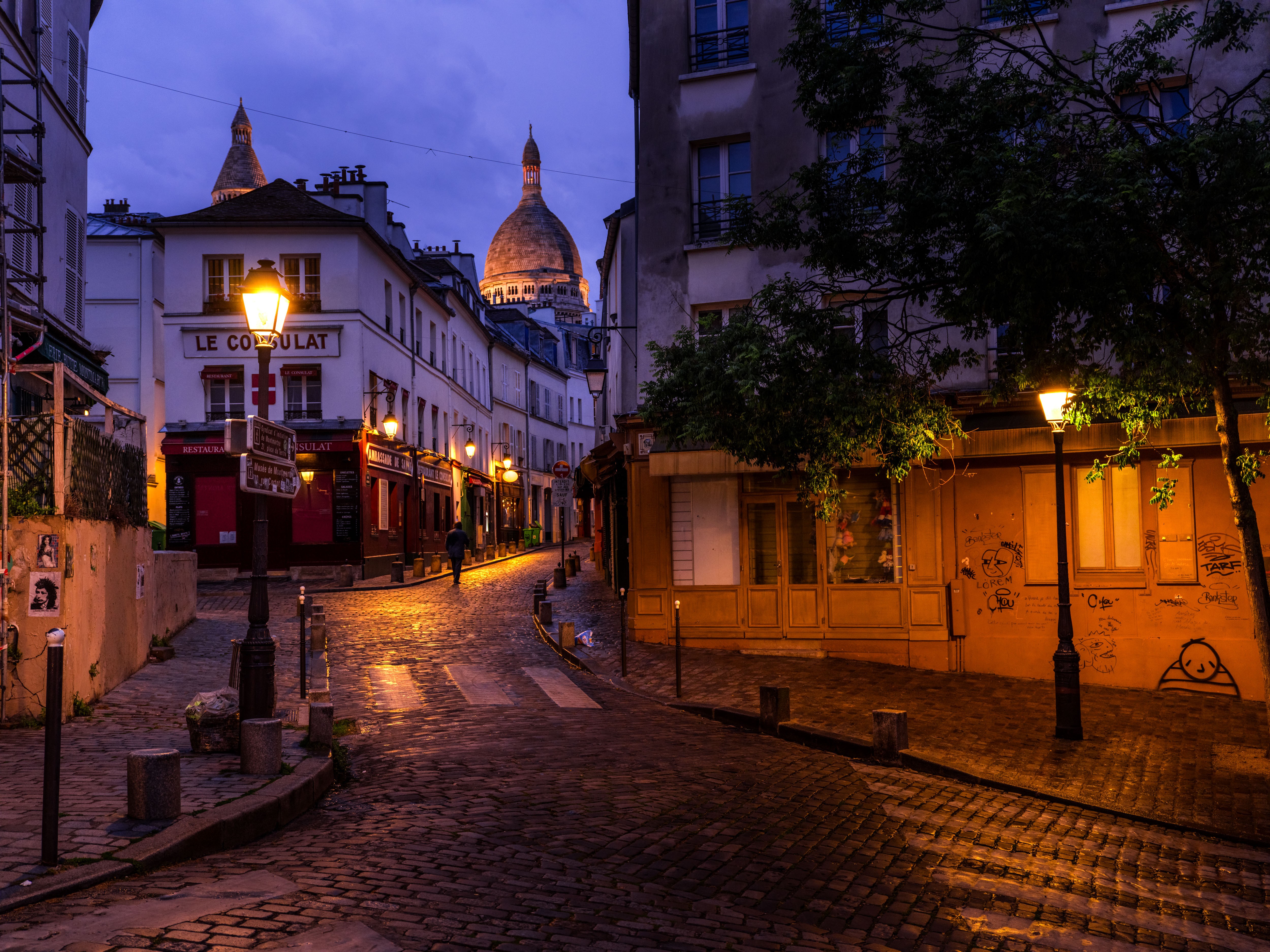 Nighttime city street with cobblestones, streetlights, buildings, and a domed building in the background.