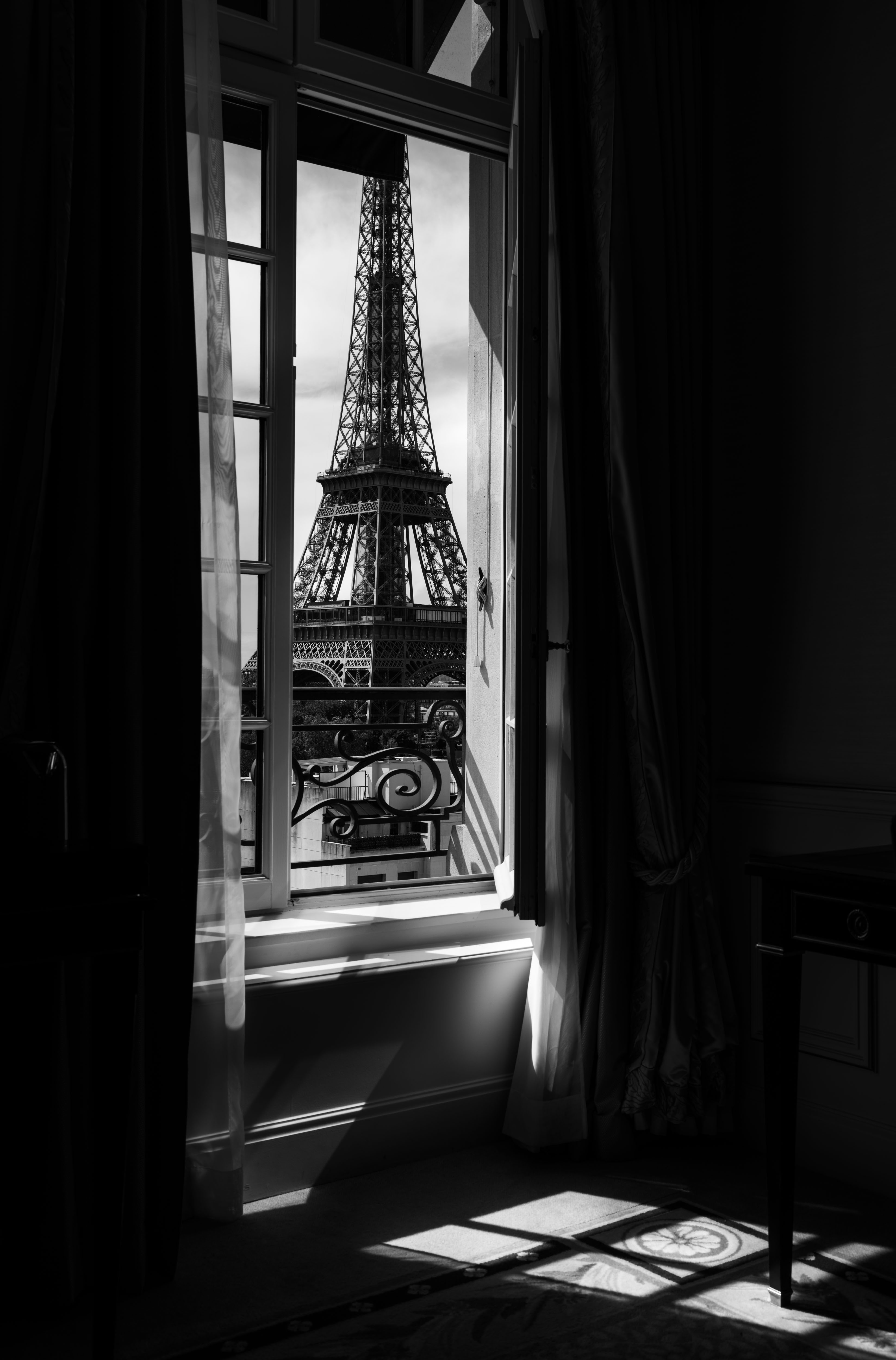View of the Eiffel Tower through an open window with curtains, sunlight casting shadows inside a room.