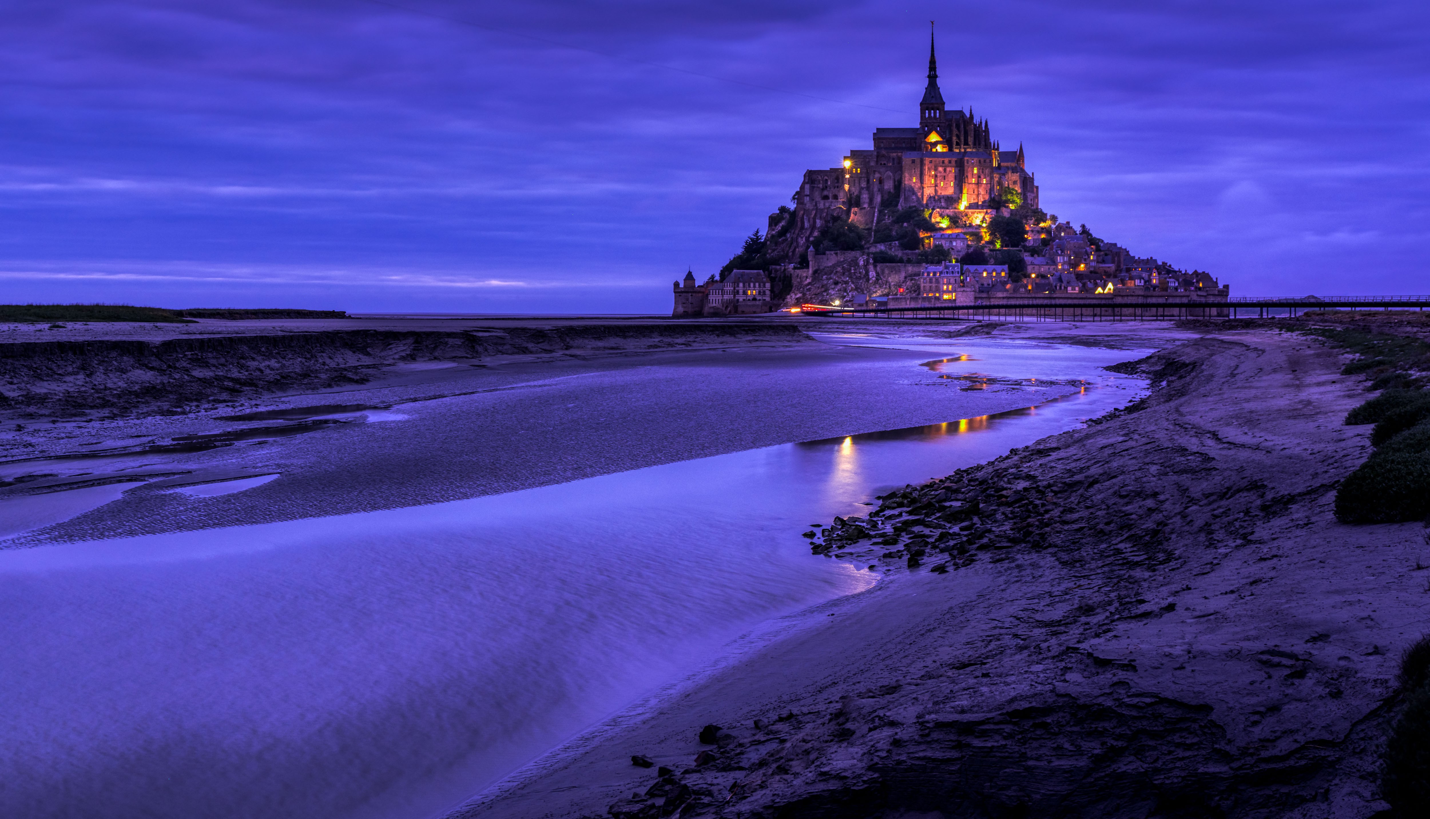 View of Mont Saint-Michel illuminated at night, with a reflective water surface in the foreground.