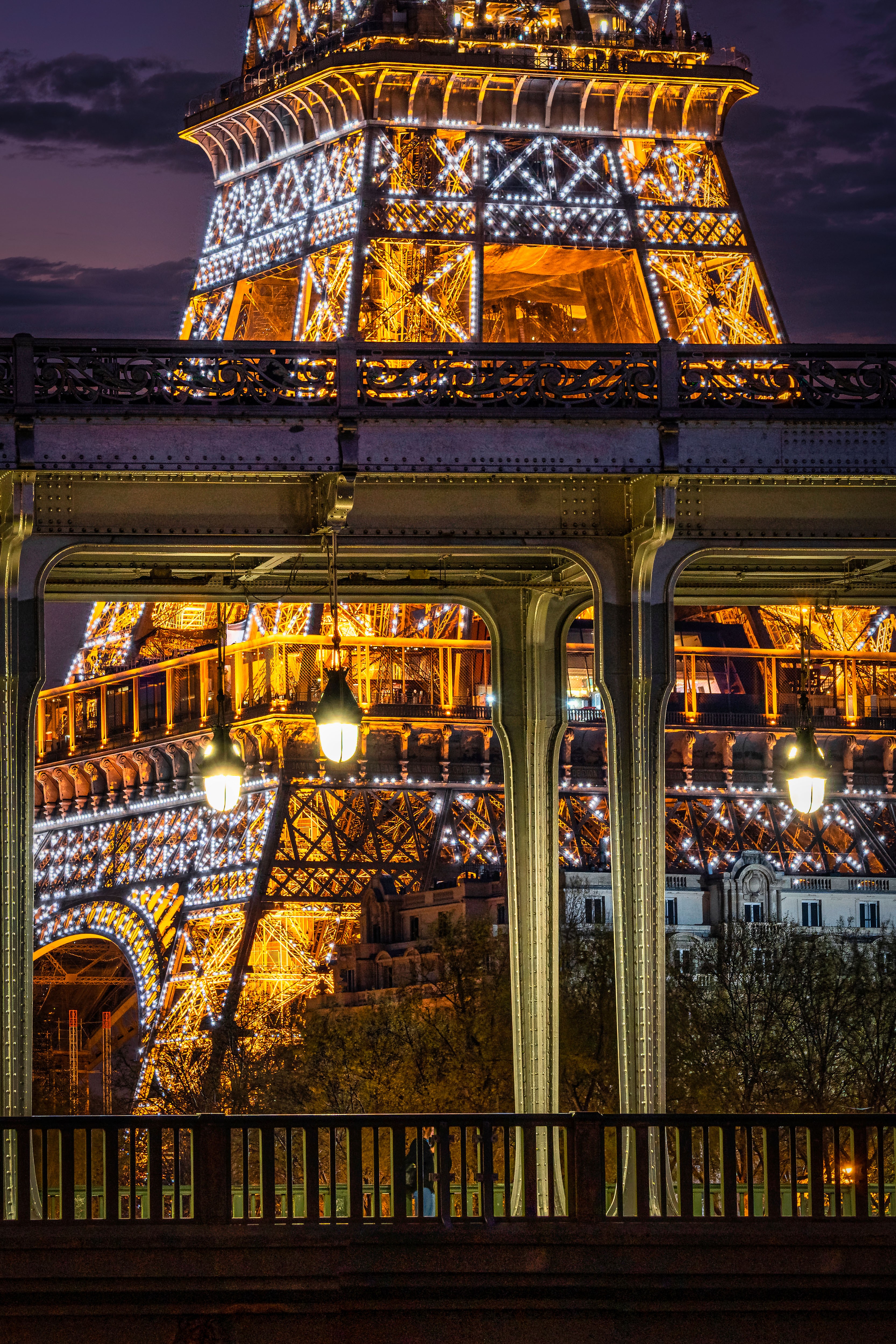Night view of the Eiffel Tower illuminated with lights, reflected in a glass window, with a bridge in foreground.