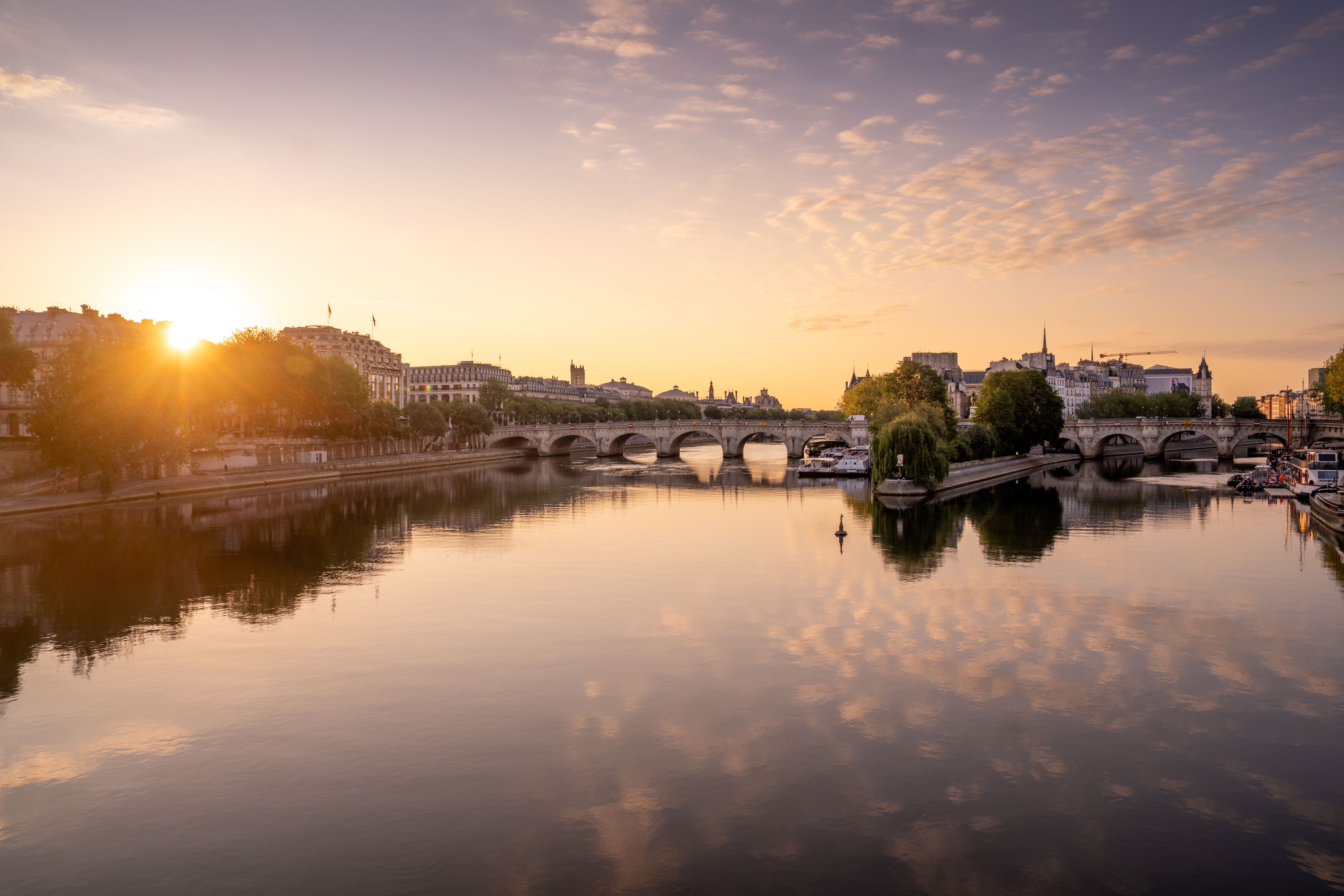 Sunset over a river with a bridge and cityscape in the background, reflected in calm water.
