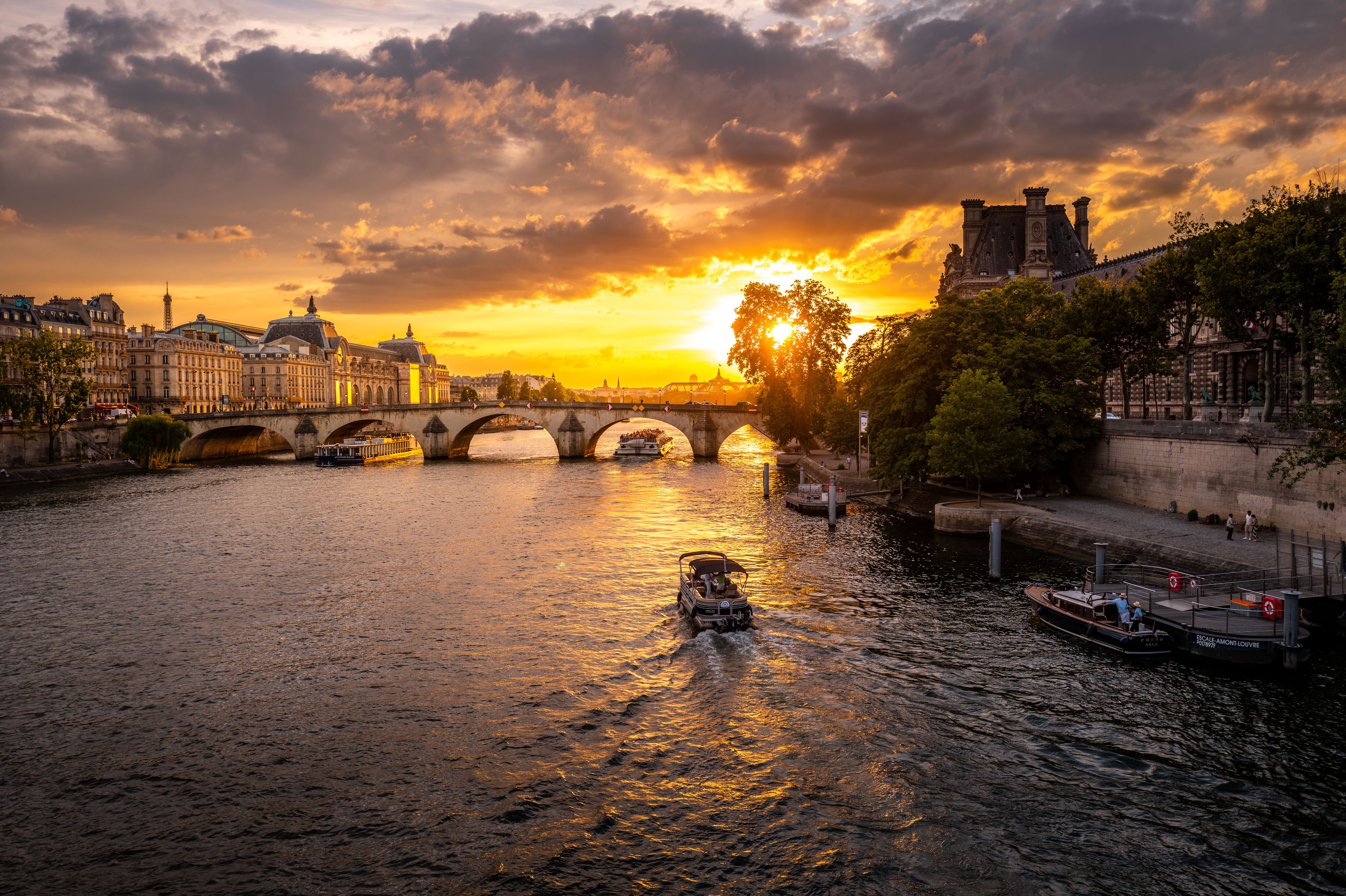 Sunset over a river with a bridge, buildings, and trees, with a boat on the water and a cloudy sky.