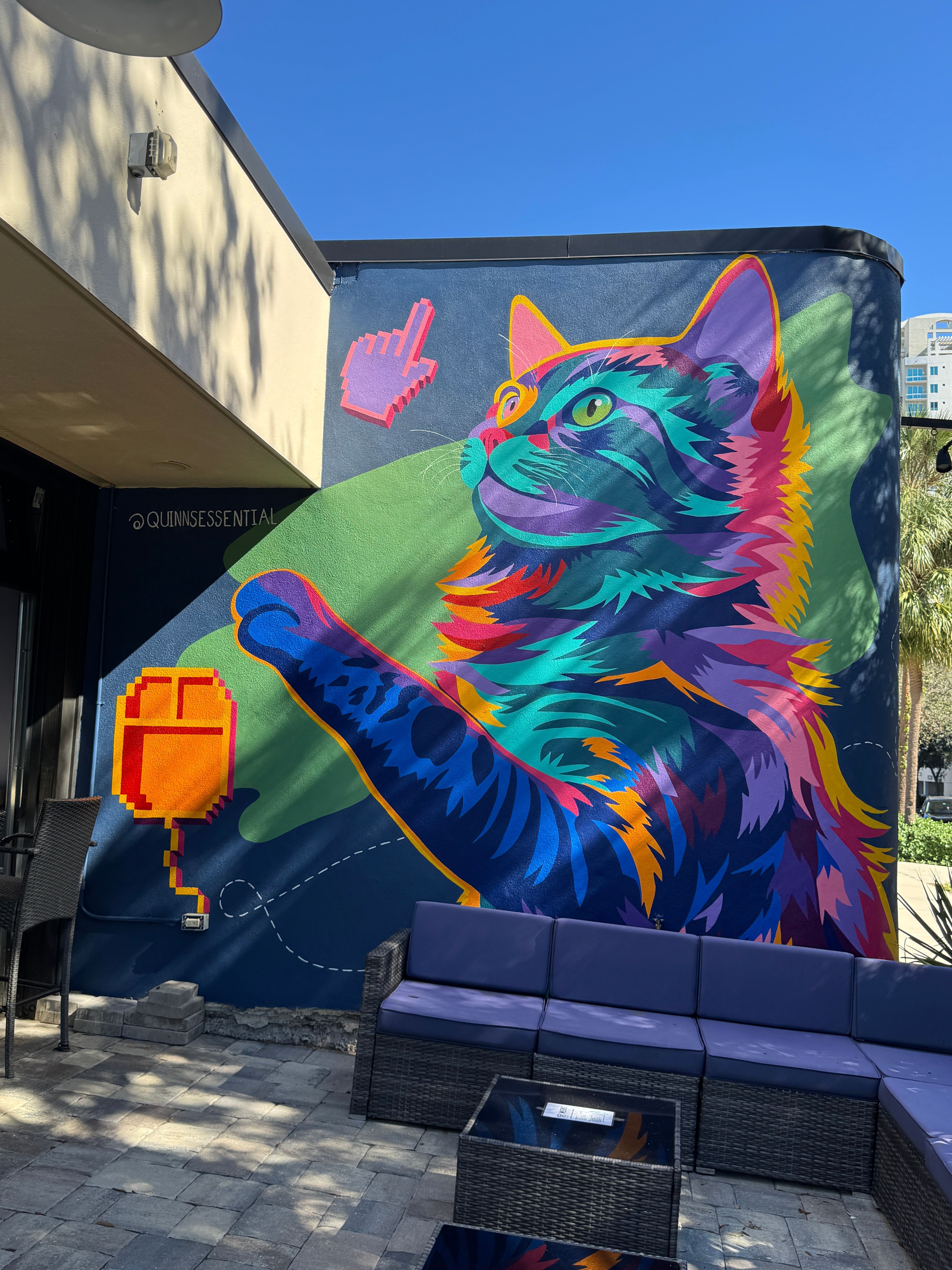 Colorful mural of a cat with vibrant blue, purple, pink, and orange fur, on a wall with a small pink hand symbol.
