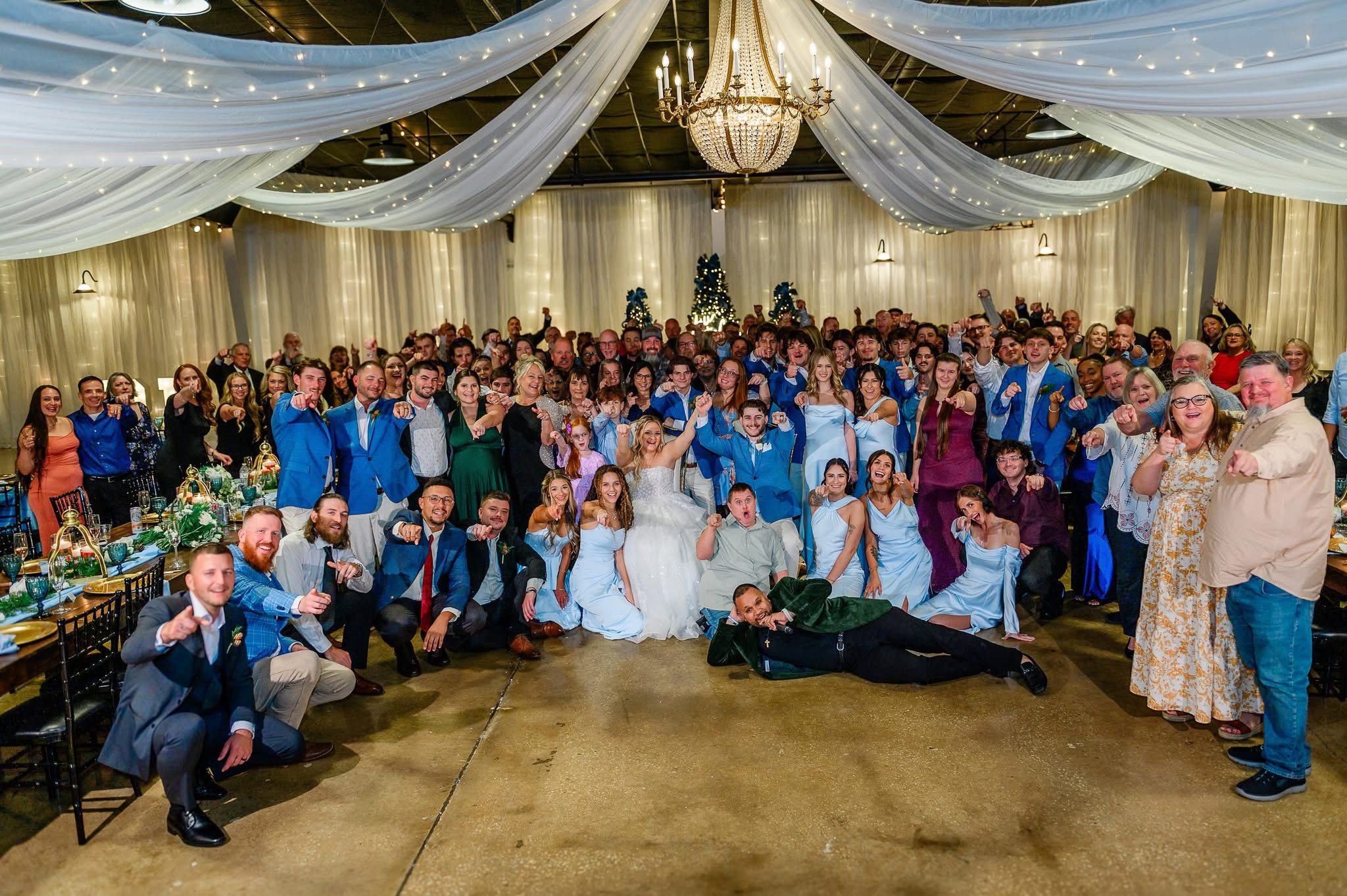 Large group of people at a wedding reception, with decorated ceiling and chandelier, some sitting and others standing, smiling.