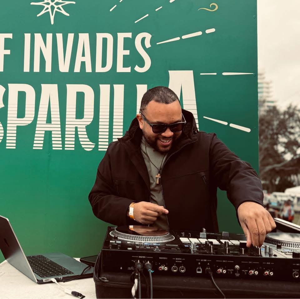 Man with sunglasses and beard operating DJ equipment outdoors, smiling, with a green sign in the background.