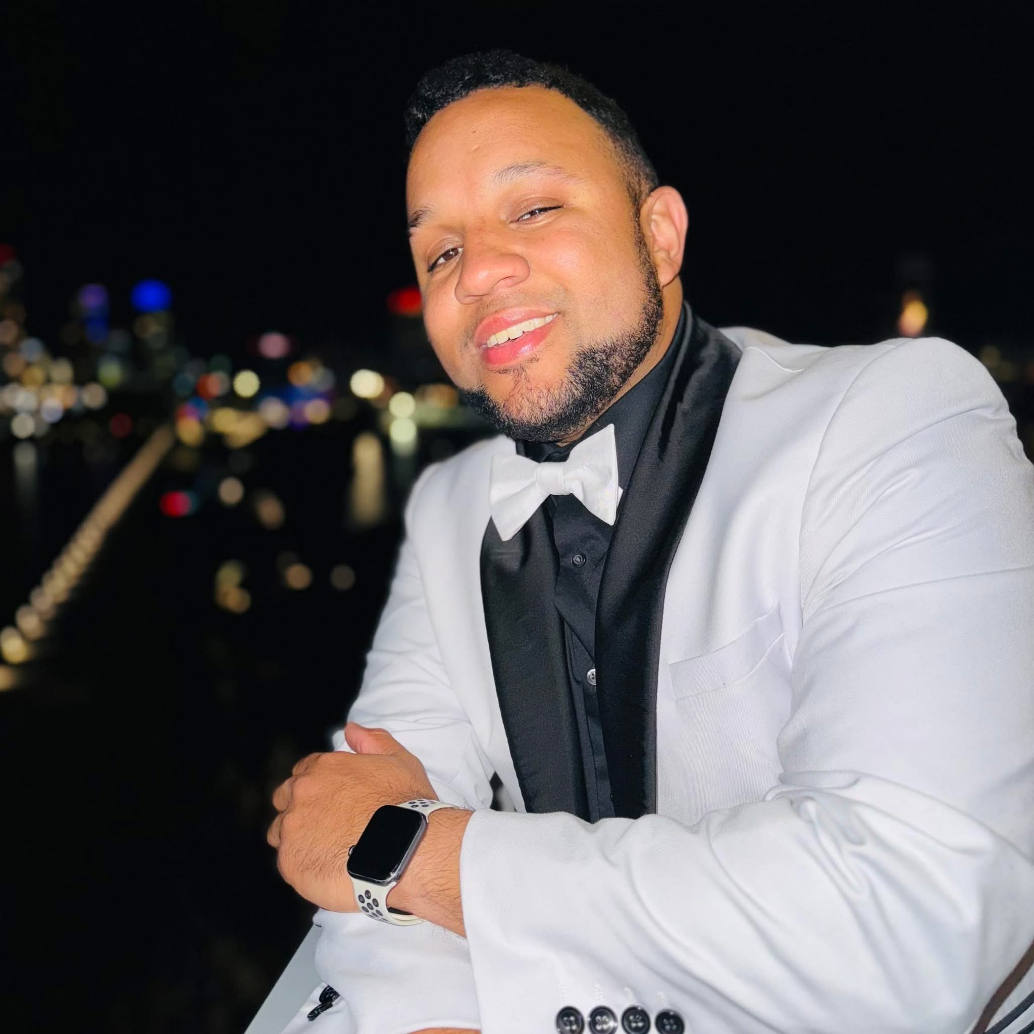 Man in white tuxedo with black bow tie smiling at night with city lights in background.