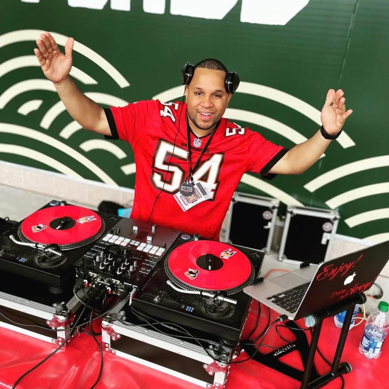 DJ wearing a red sports jersey with arms raised, standing behind turntables and a laptop, in a green and white background.