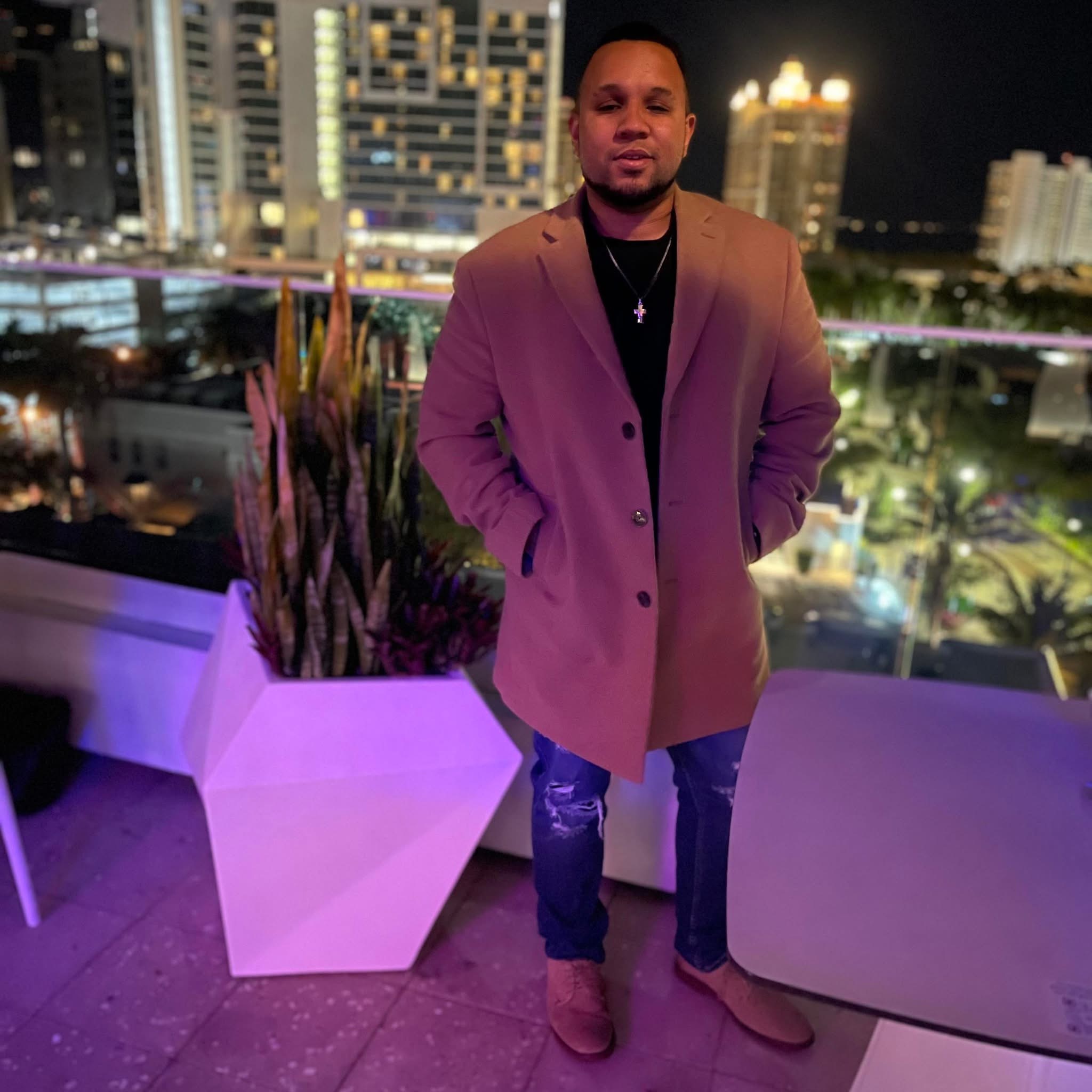 Man standing outdoors at night with city lights in background, wearing a beige coat and jeans, hands in pockets.