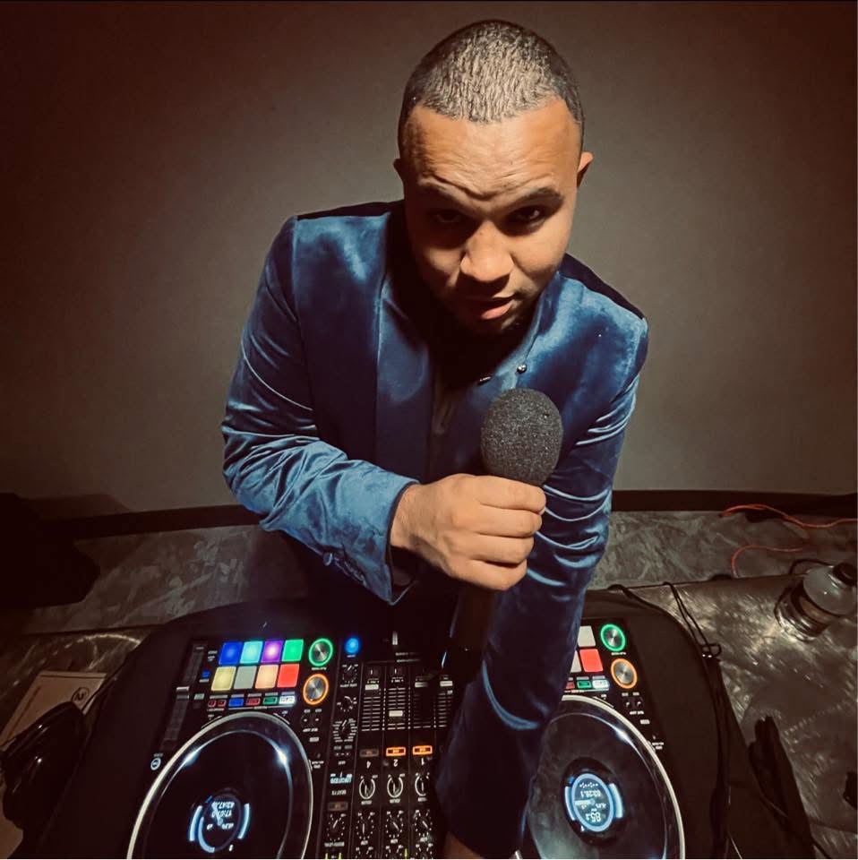 Person in a blue jacket holding a microphone, standing behind DJ equipment, looking at the camera.