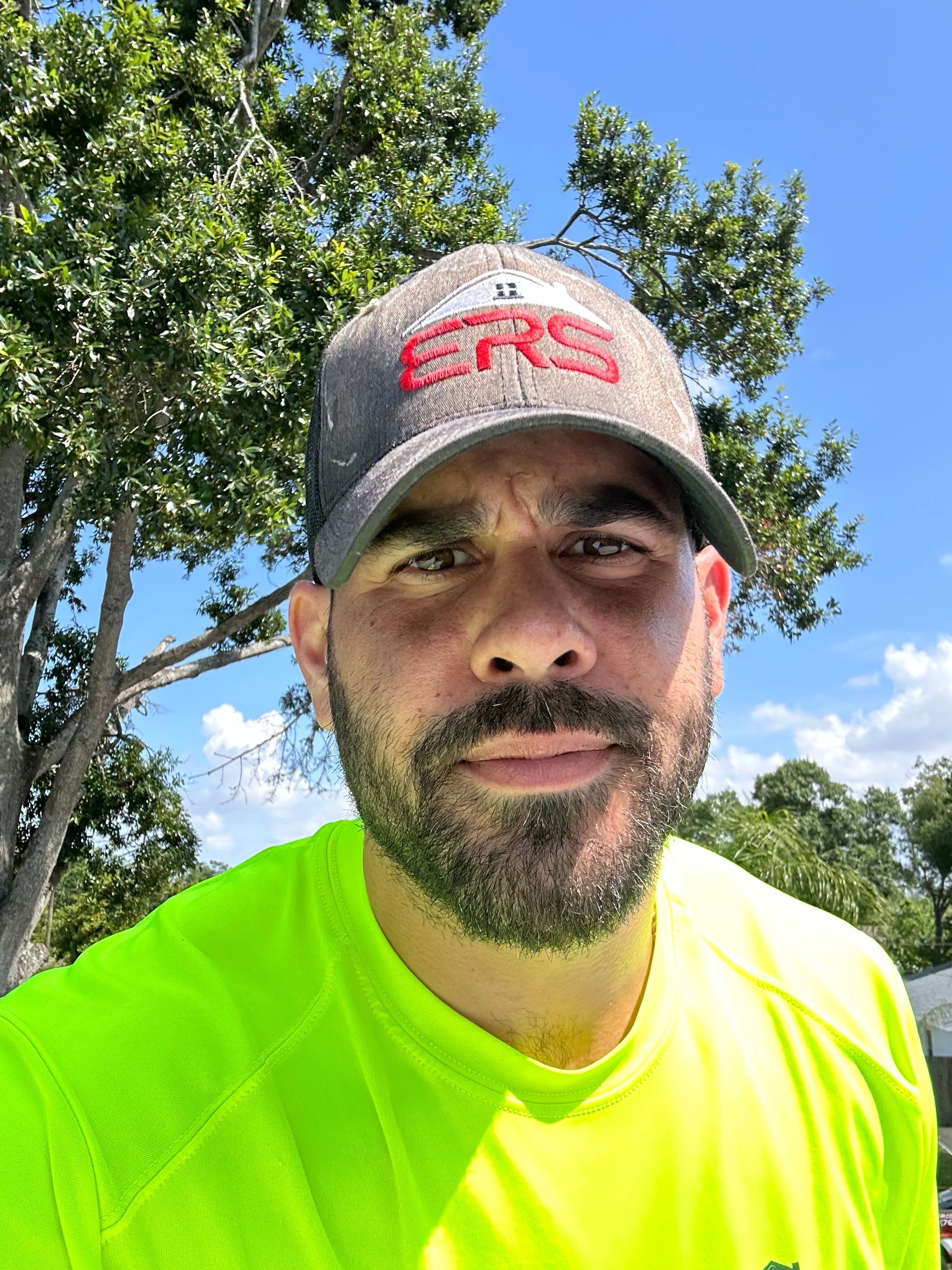 Man with a beard wearing a gray cap and bright yellow shirt outdoors, trees and blue sky in background.