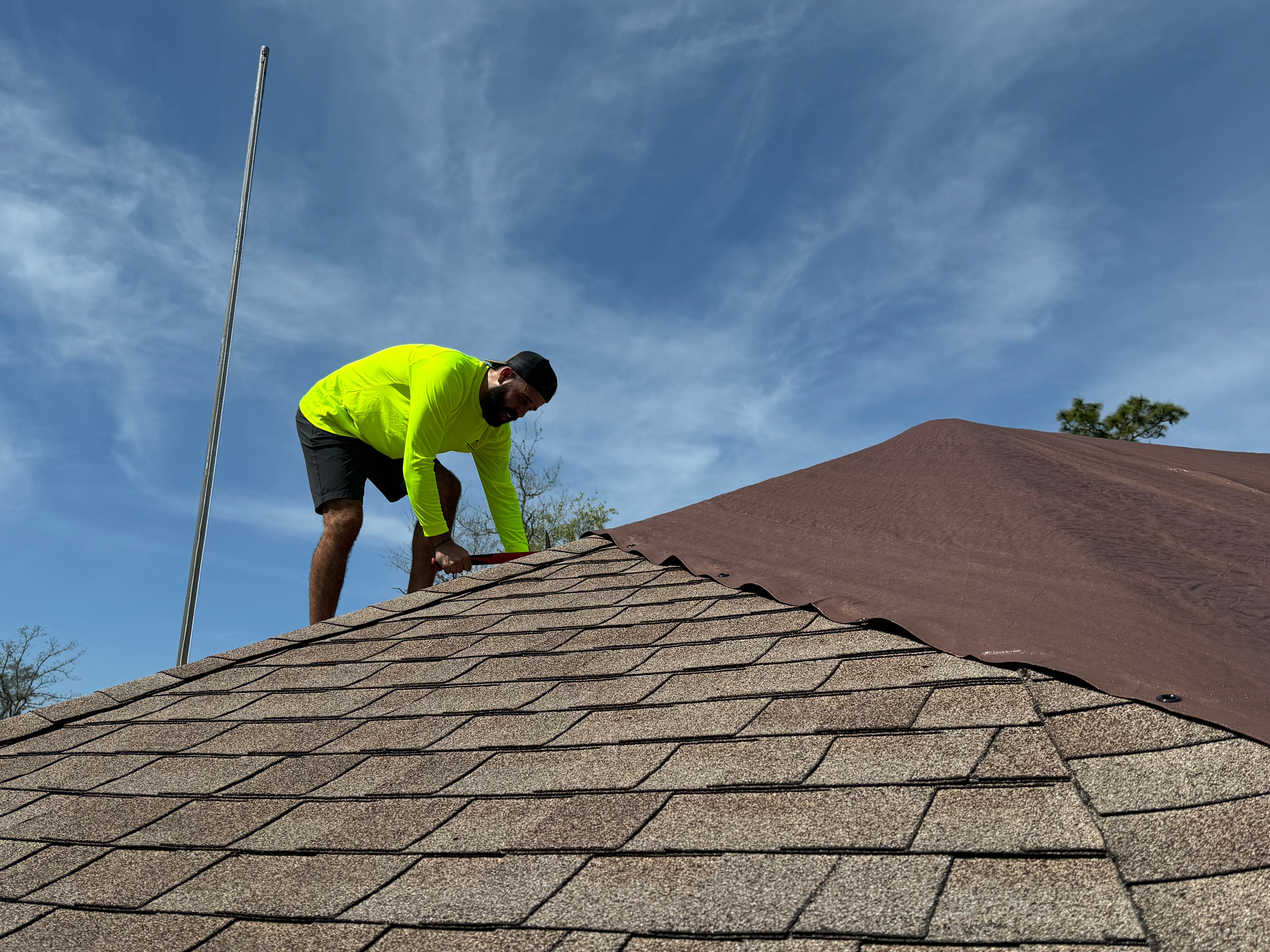 Person in a bright yellow jacket working on a roof with shingles, under a blue sky with clouds.