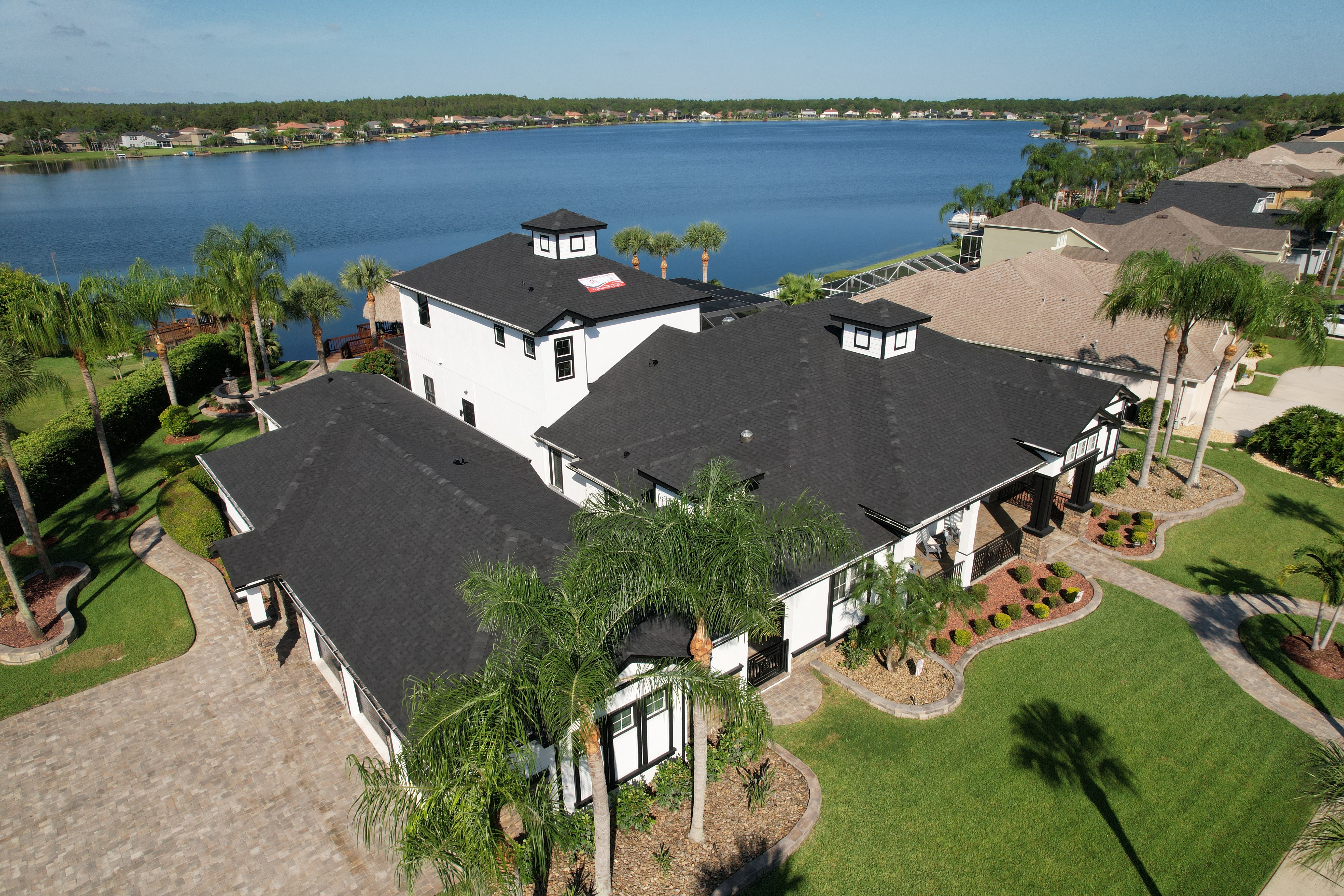A large white house with a black roof near a body of water, surrounded by green lawns and palm trees.