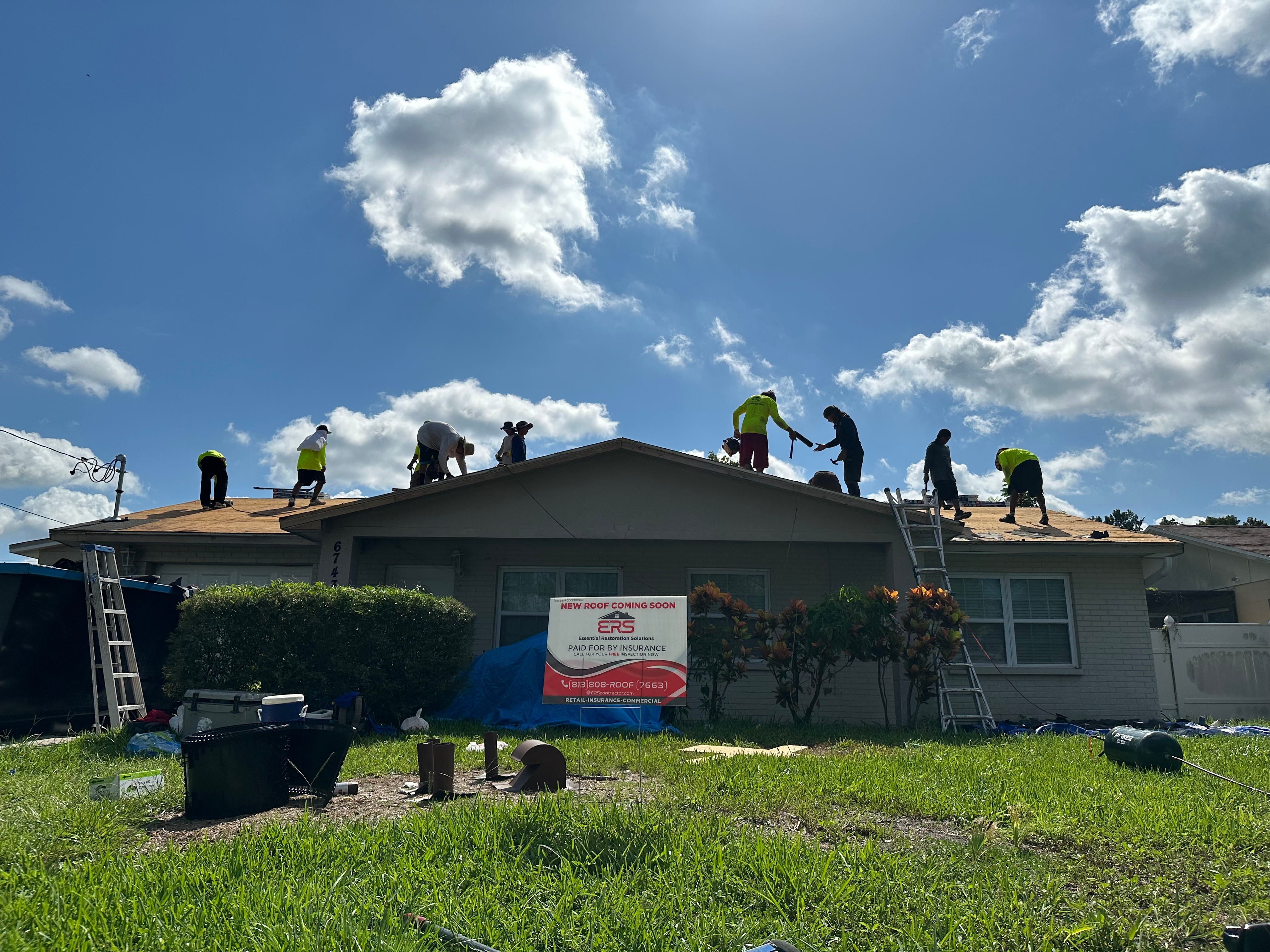 People working on a house roof under a partly cloudy sky, with tools and equipment on the ground.