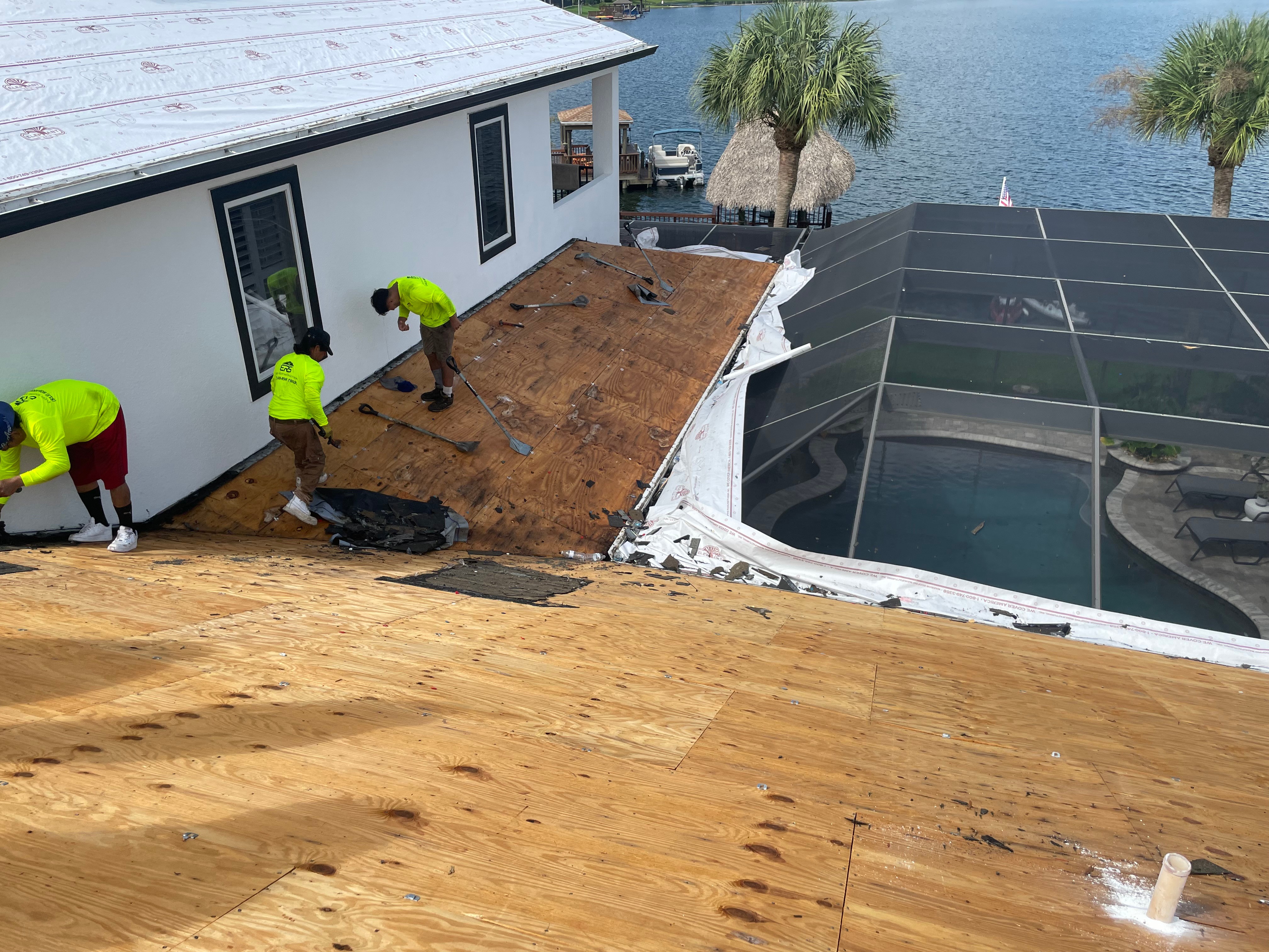 Workers in bright yellow shirts are installing a wooden deck outside a house near water, with palm trees and a pool nearby.