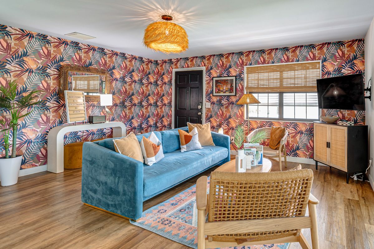 Living room with colorful tropical wallpaper, blue sofa, rattan armchair, wooden furniture, and a window with blinds.