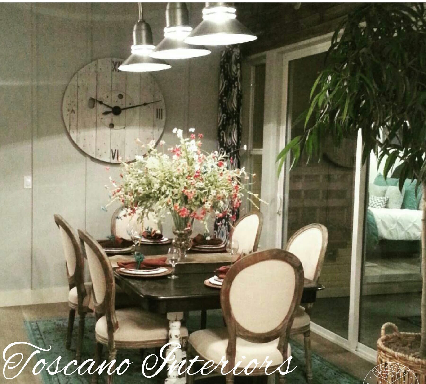 Dining table with six chairs, floral centerpiece, wall clock, and hanging lights in a modern room with sliding glass door.
