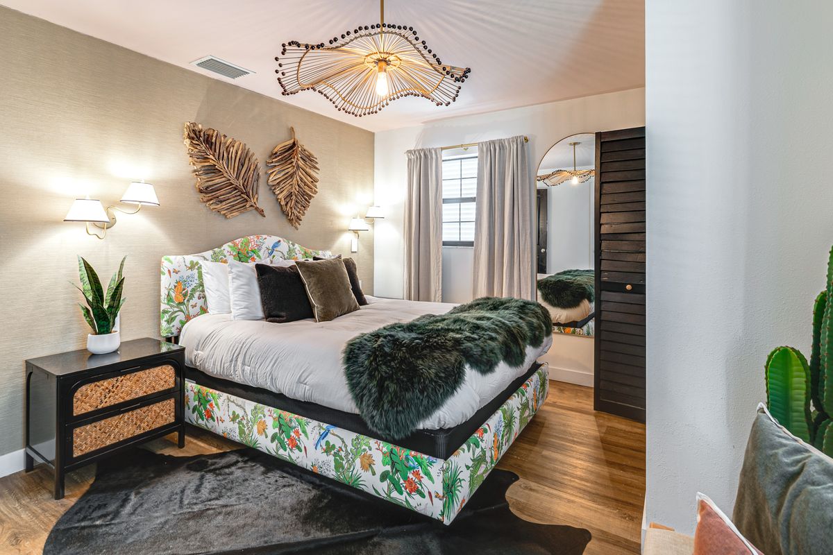 Bedroom with bed, floral bed frame, black nightstand, potted plant, wall decor, window with curtains, and wooden floor.