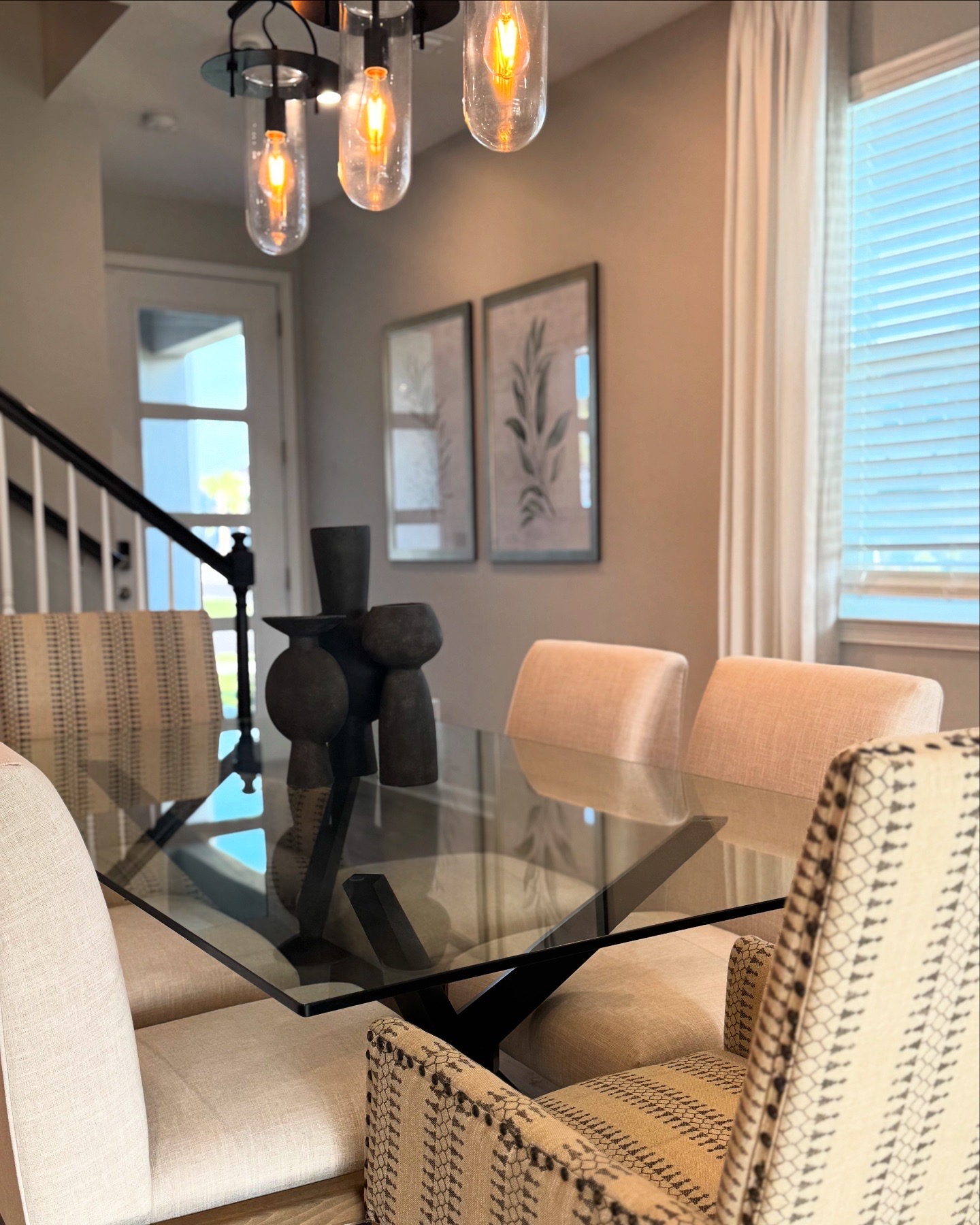 Living room with glass dining table, beige chairs, wall art, window with blinds, and hanging light fixtures.