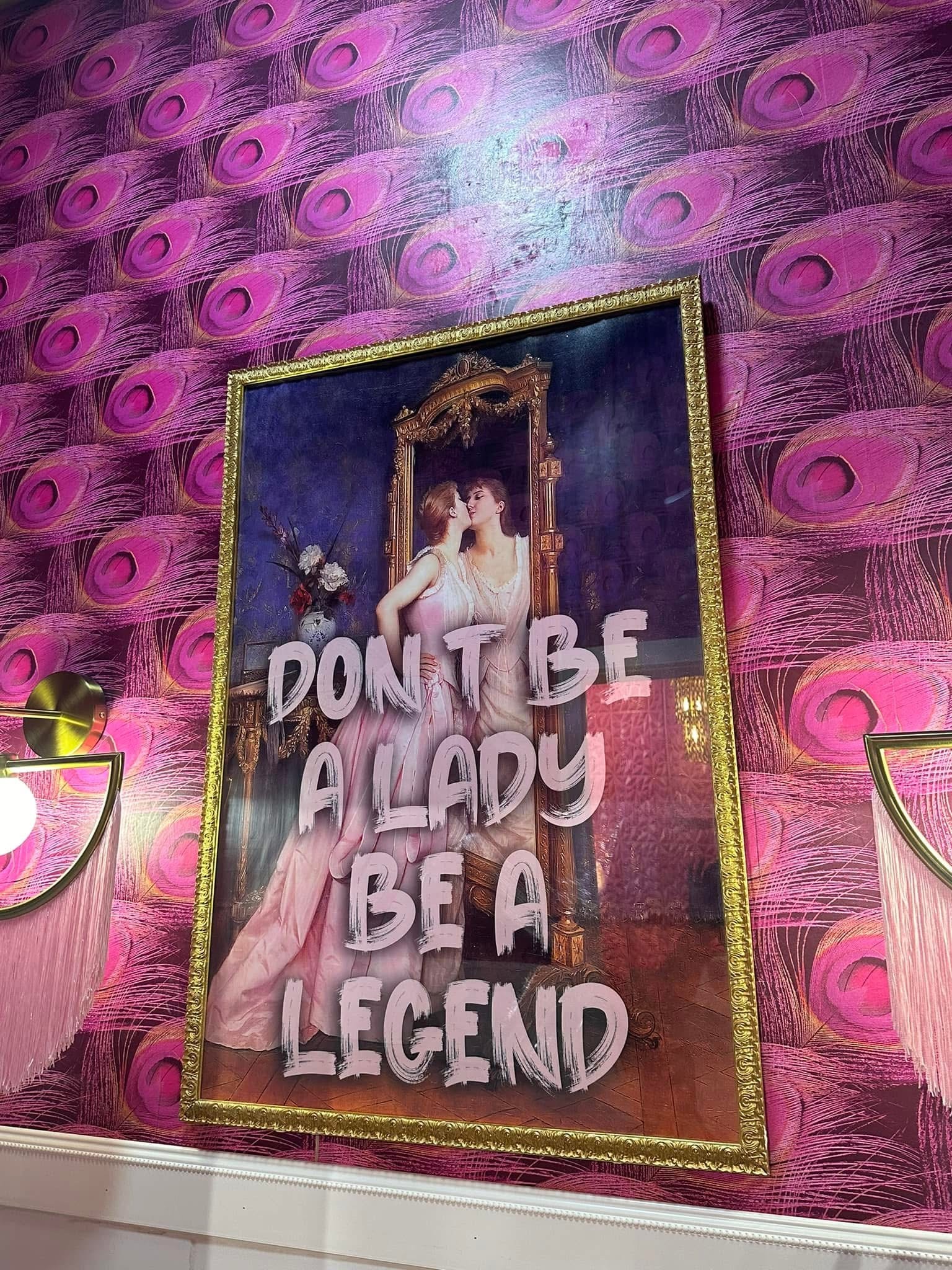 Poster with a woman in a pink dress and the text 'Don't be a lady, be a legend' on a pink patterned wall.