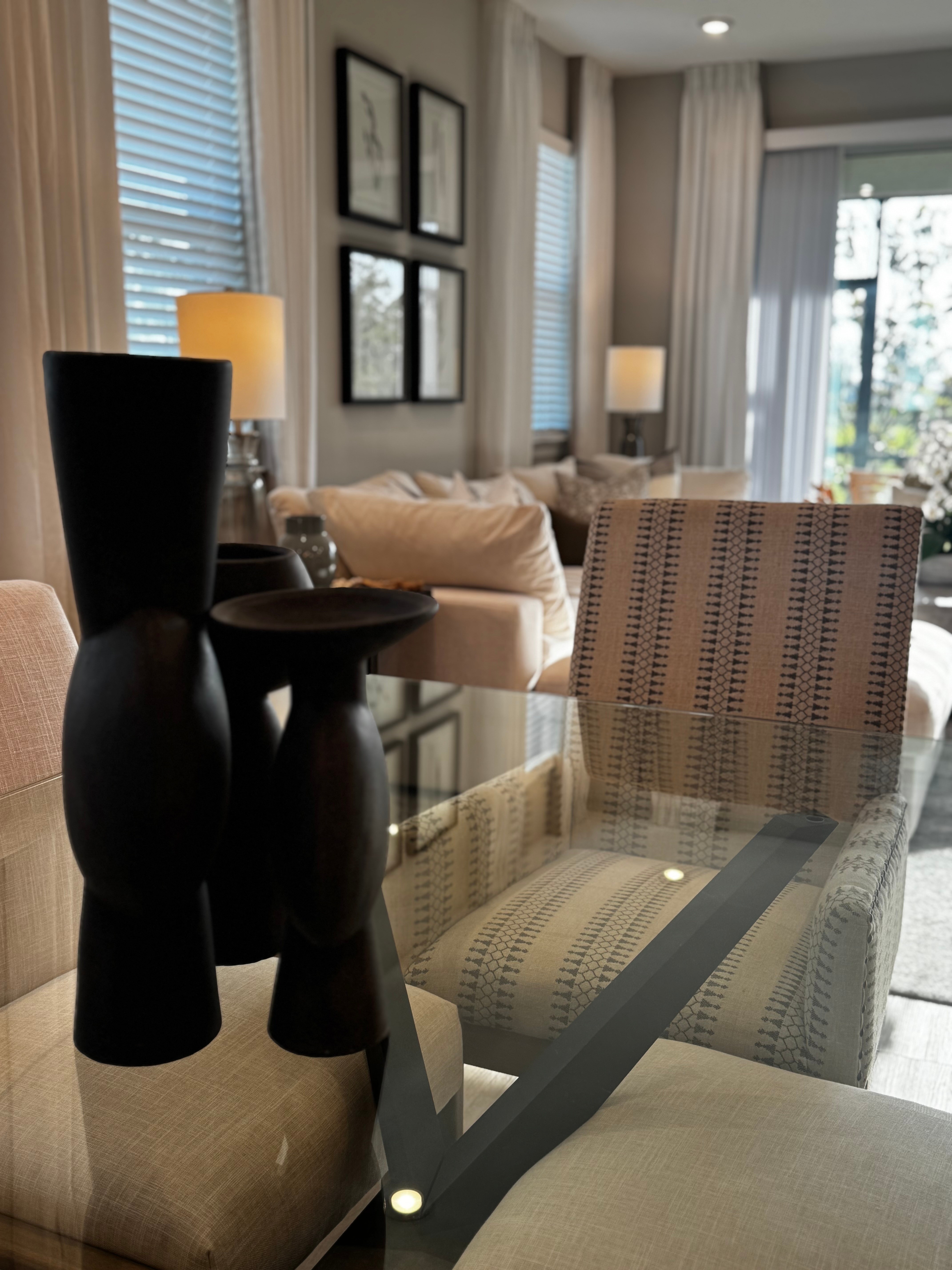 Living room with beige sofa, patterned armchair, glass table, and wall art, illuminated by natural light and lamps.