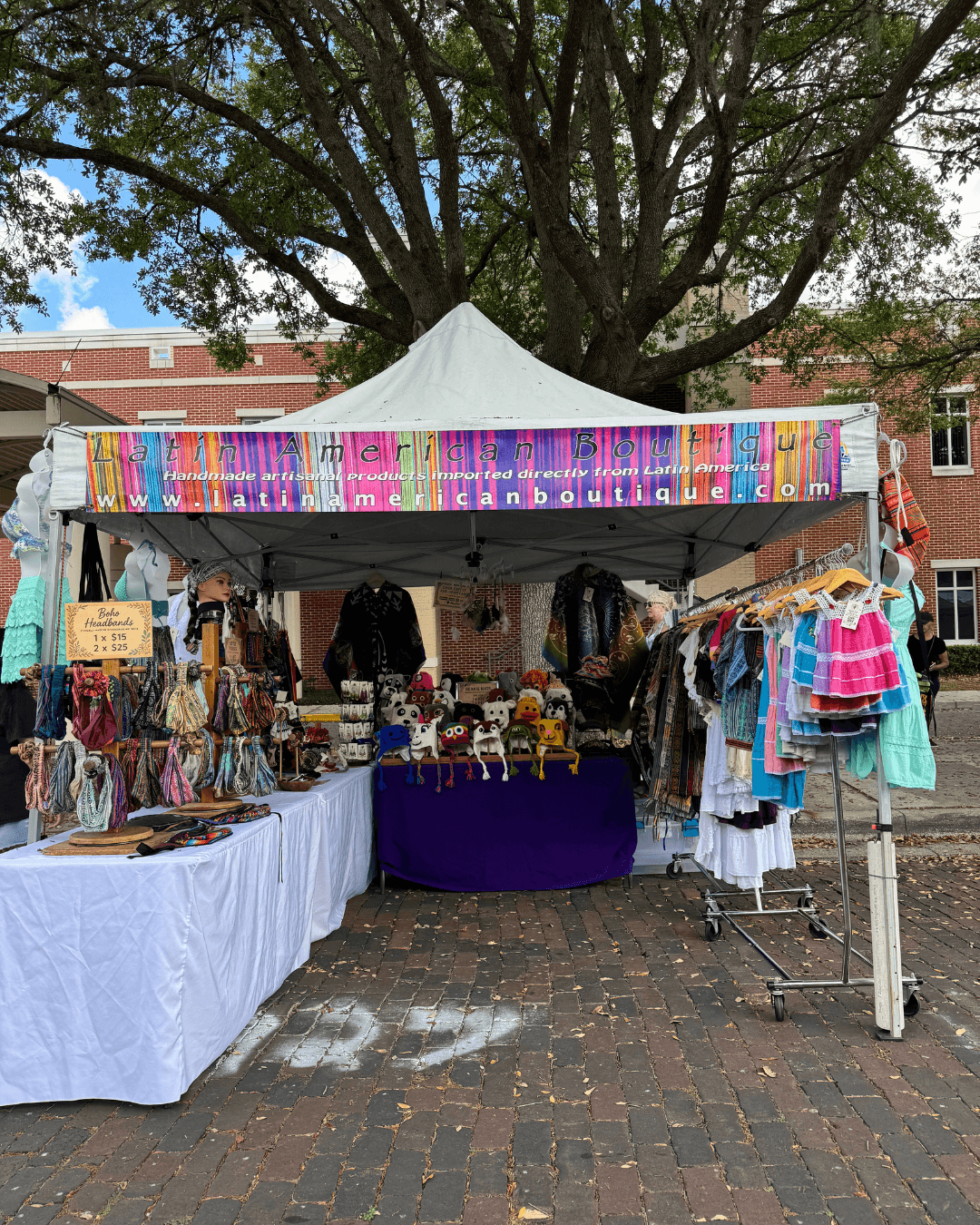 Outdoor market stall with clothing and accessories under a white canopy, set on brick pavement with a large tree in background.