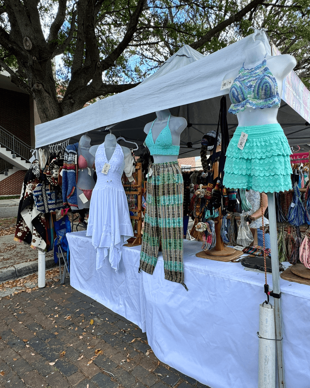 Clothing stall with mannequins displaying dresses and pants, surrounded by accessories and other clothing items, under a white canopy.
