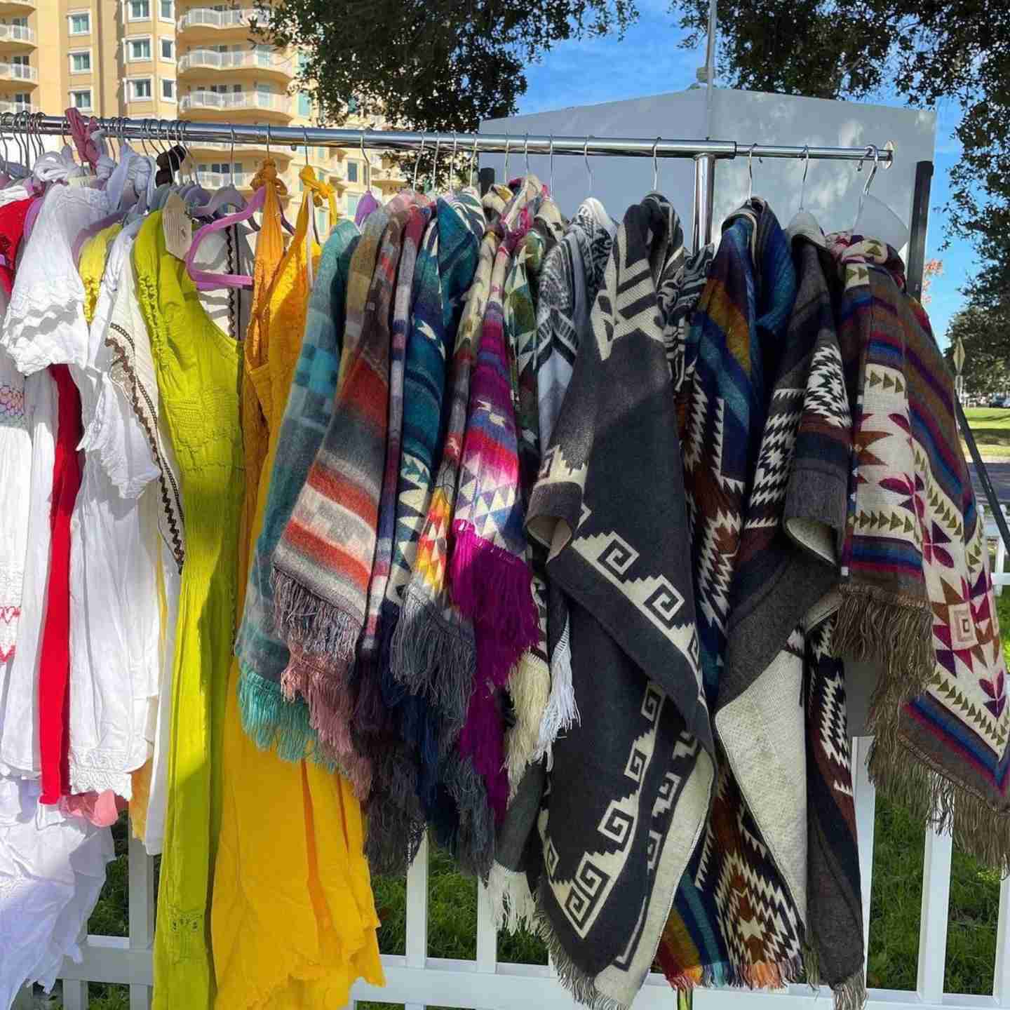 Clothing rack with colorful scarves and garments outdoors, trees and buildings in background.
