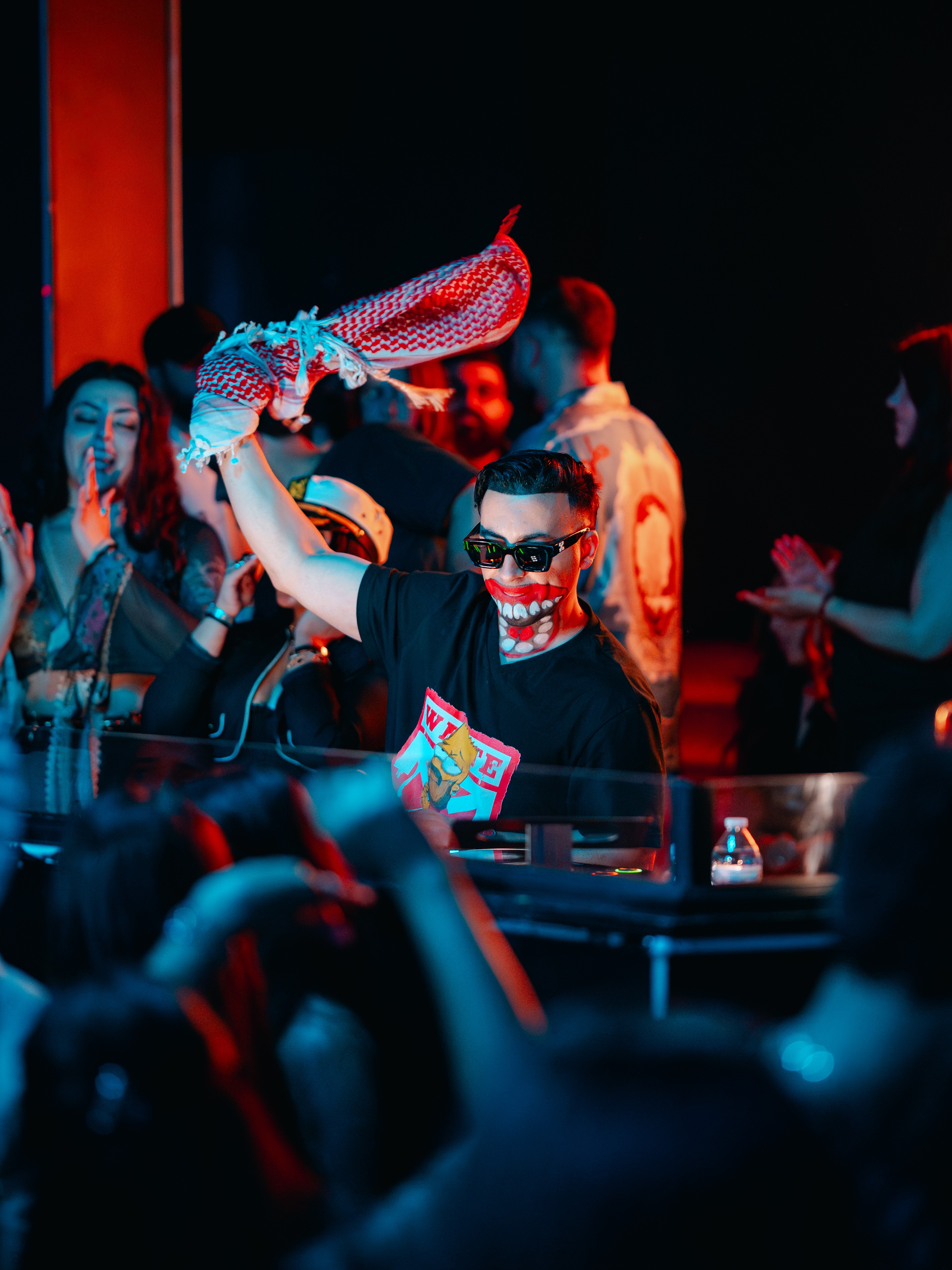 Person wearing sunglasses and a mask, holding a fish-shaped object, surrounded by a crowd in a dark setting.